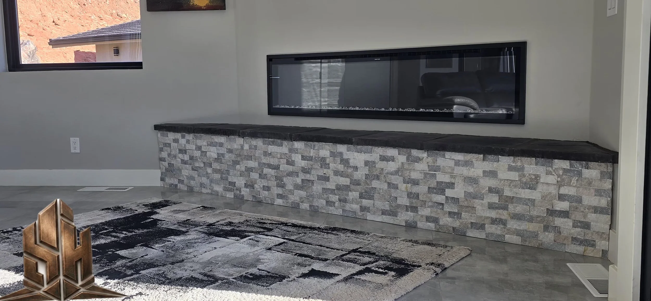 Living room with fireplace built into a stone wall, gray tile floor, and a large window showing an outdoor view.