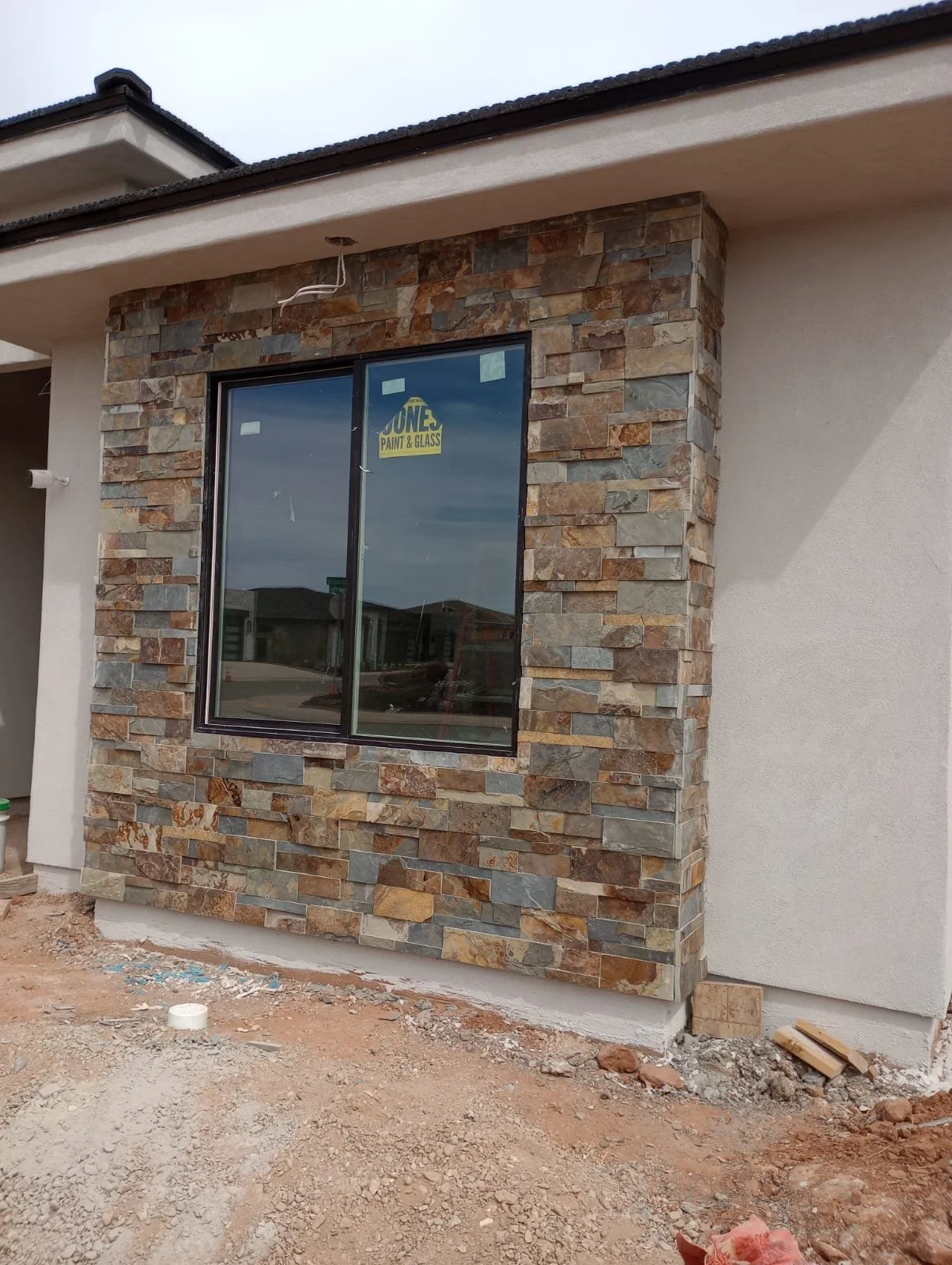 Under construction house exterior with a large window surrounded by stone veneer and a concrete wall, dirt ground, and construction materials nearby.