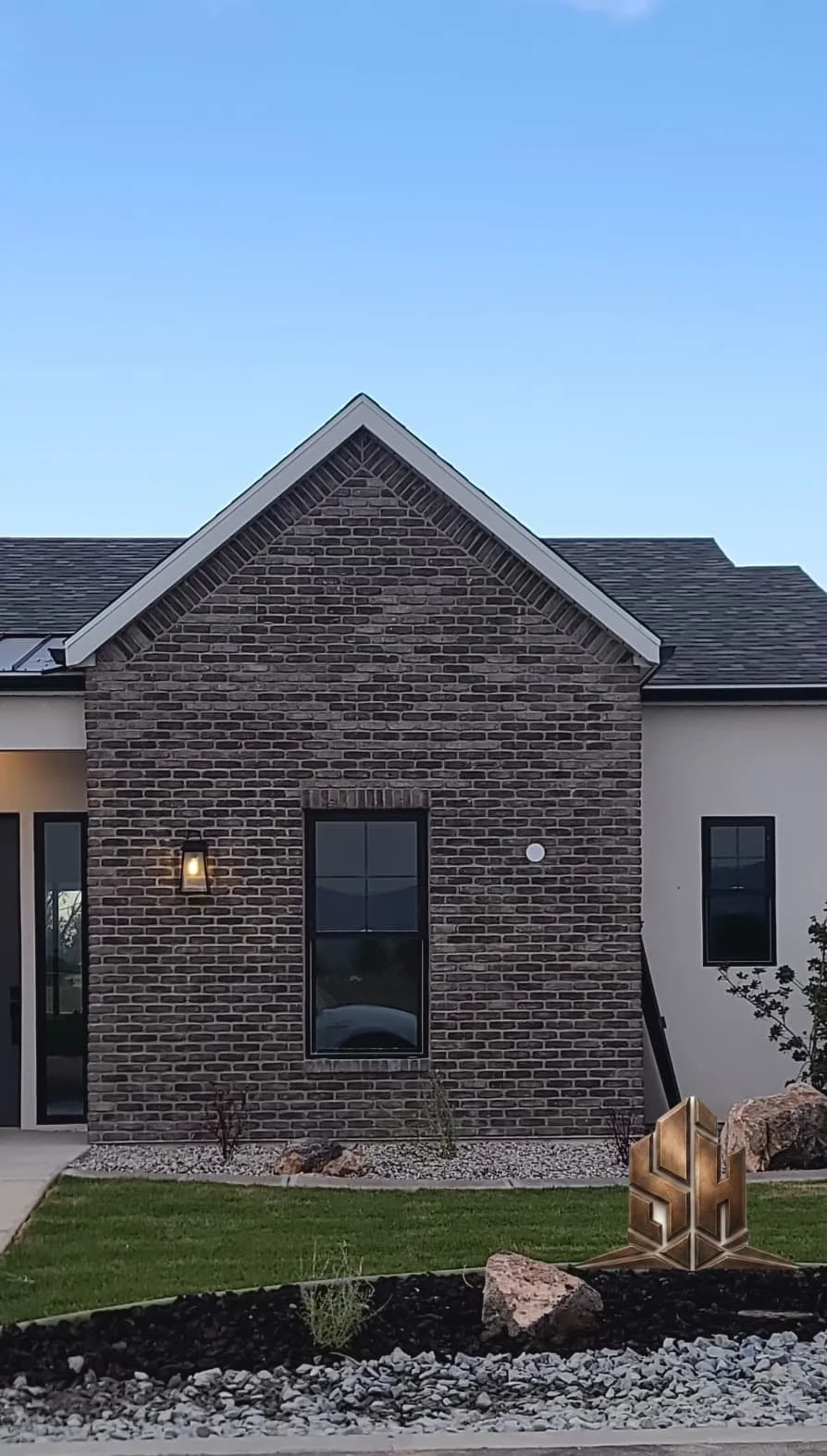A modern house with brick and white exterior, a gable roof, a large window, and minimal landscaping with rocks and a small plant in the front yard.