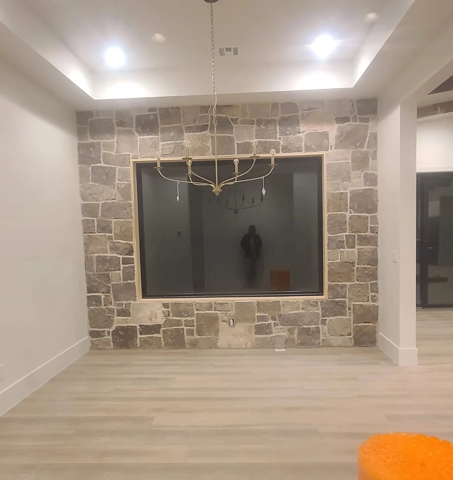 Interior of a room with a stone accent wall, a chandelier hanging from the ceiling, and a large mirror reflecting the room. There is a small orange cushioned stool in the bottom right corner and wooden flooring.