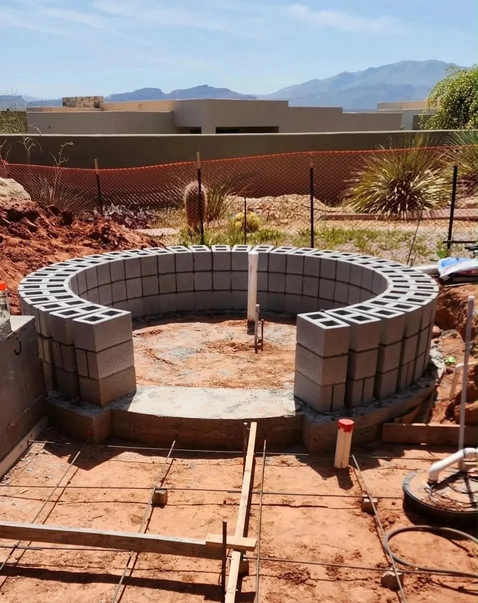 Construction site with circular block wall, outdoor setting with mountains in the background, and construction materials and tools in the foreground.