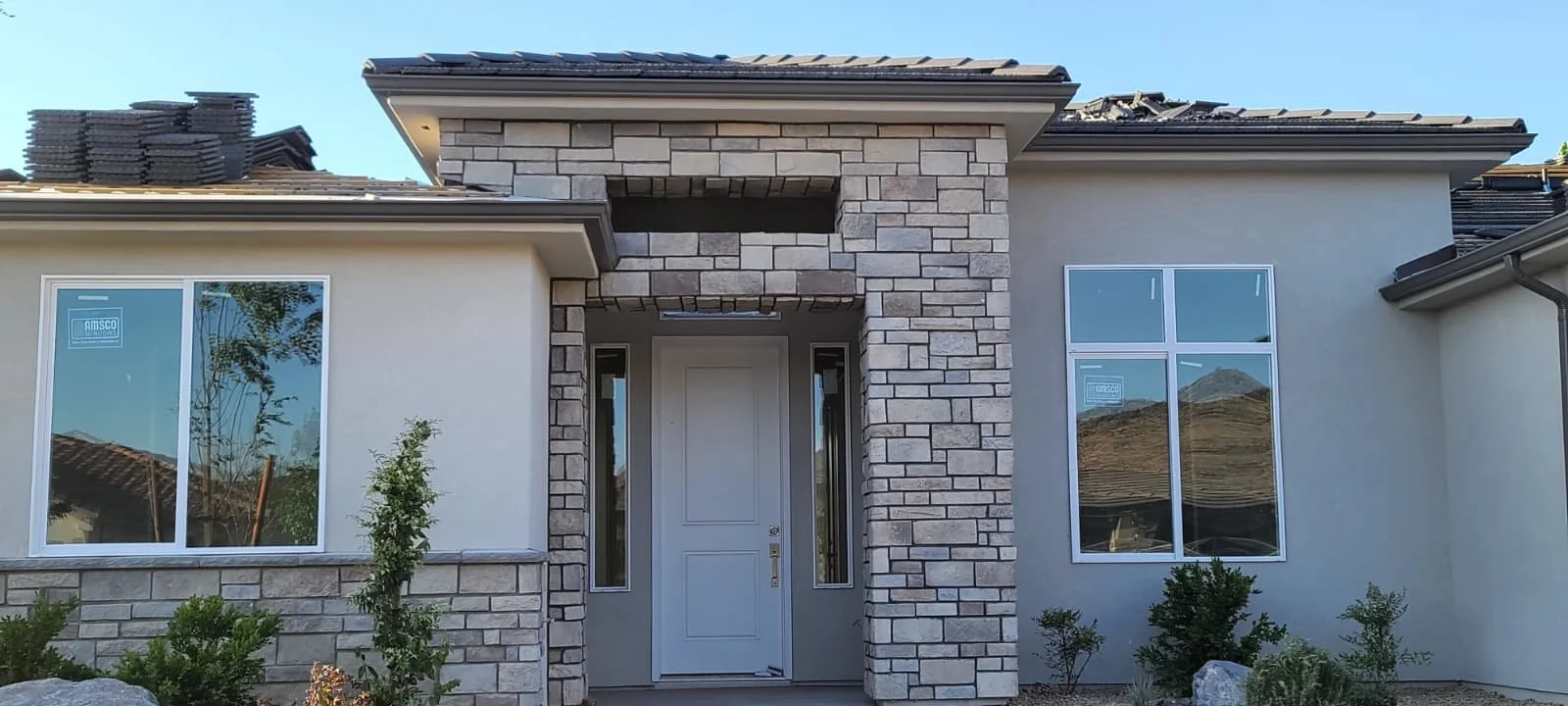 Newly constructed house with light-colored walls, stone accents around the front door, large windows, and a landscaped front yard with small bushes and rocks.