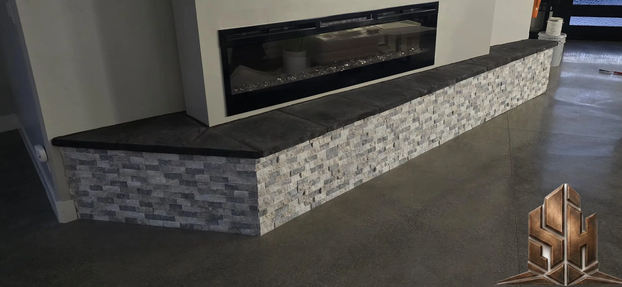A modern fireplace with a glass front, set into a white wall, above a low stone wall made of stacked rectangular bricks, with a dark stone top, in a room with a polished concrete floor.