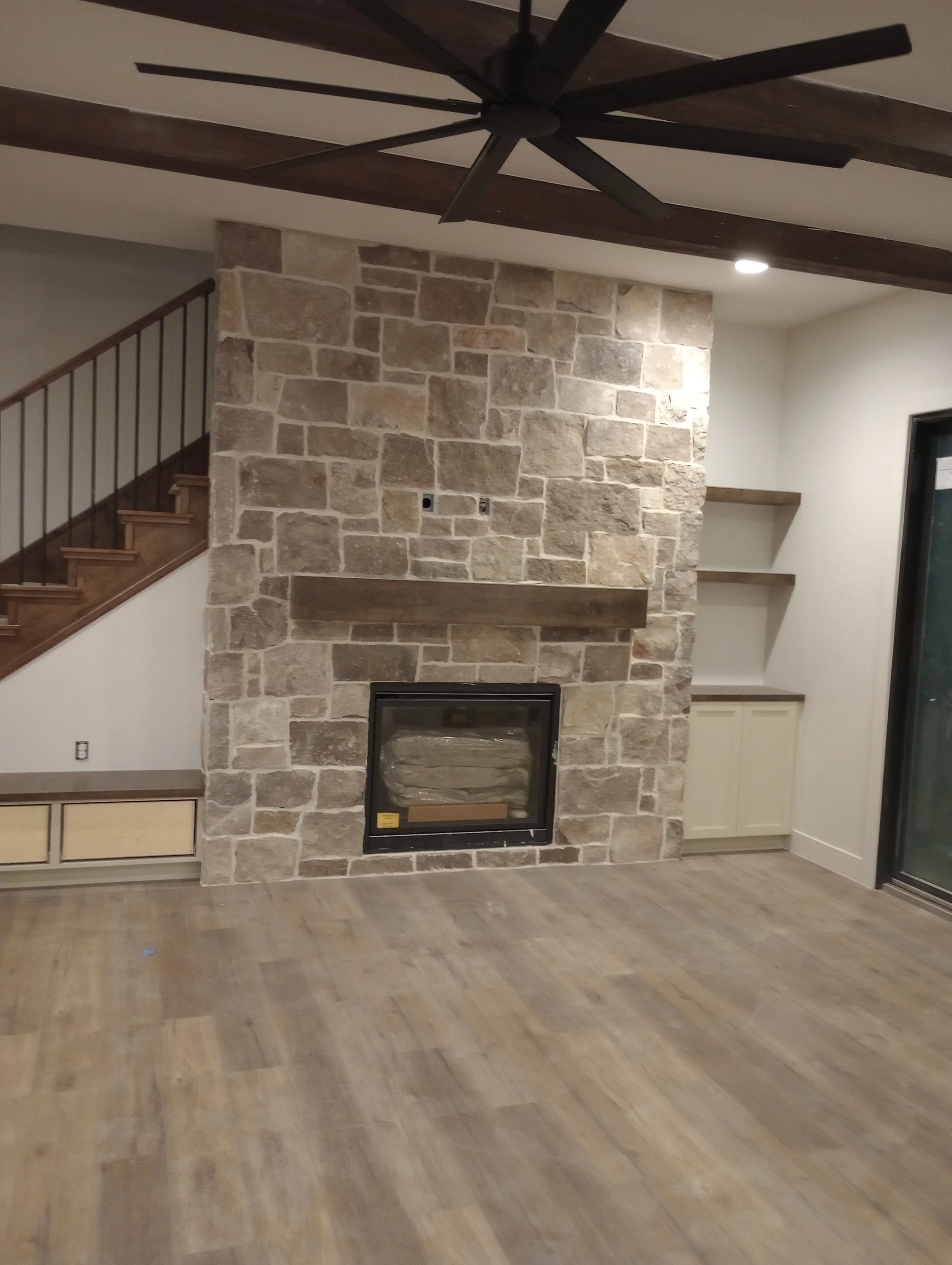 Interior room featuring a stone fireplace with a wooden mantel and a black fireplace insert. Adjacent to the fireplace are two open shelves, a white cabinet below, and a staircase with a wooden handrail on the left. The ceiling has dark wooden beams and a large black ceiling fan. The room has wood-look flooring and a sliding glass door on the right.