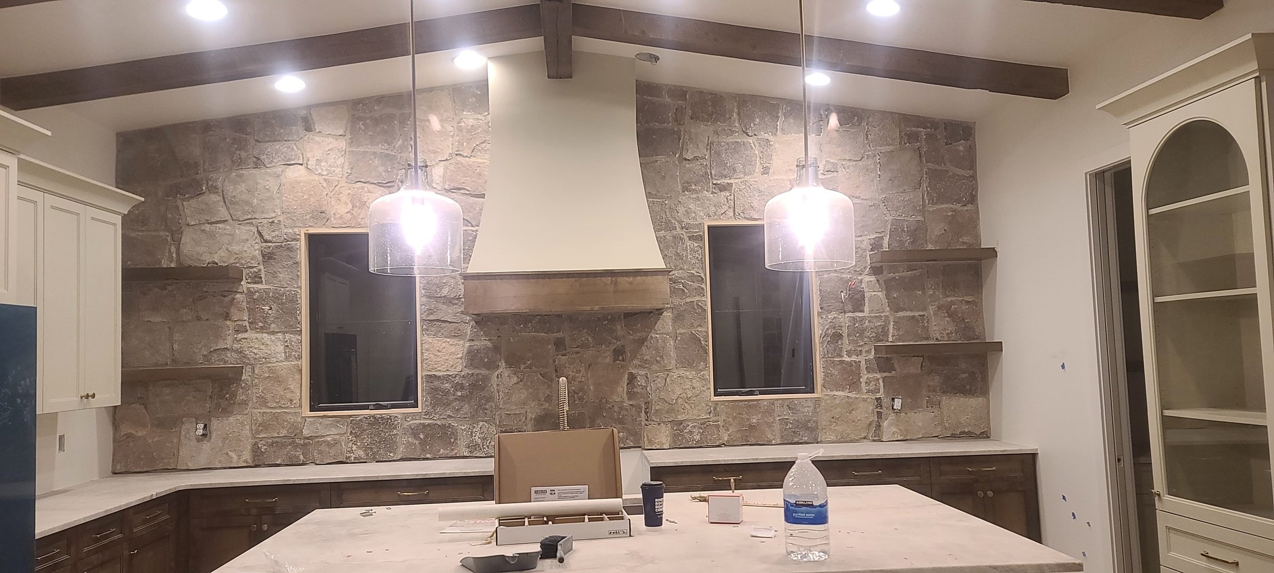 View of a kitchen with a large island in the center. The background features a stone wall with two narrow windows. Two pendant lights hang over the island. There are shelves on either side of the windows and cabinetry on the sides of the wall. The upper cabinets are cream-colored, and the lower cabinets are brown. Various items are on the island, including a water bottle, paper, and small tools.