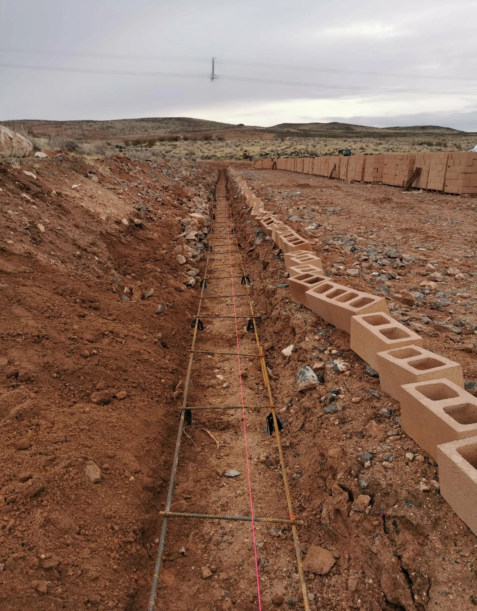 A construction site in a desert area with stacks of bricks on the right side and a trench being prepared with rebar and a pink string line for laying foundation, overcast sky in the background.