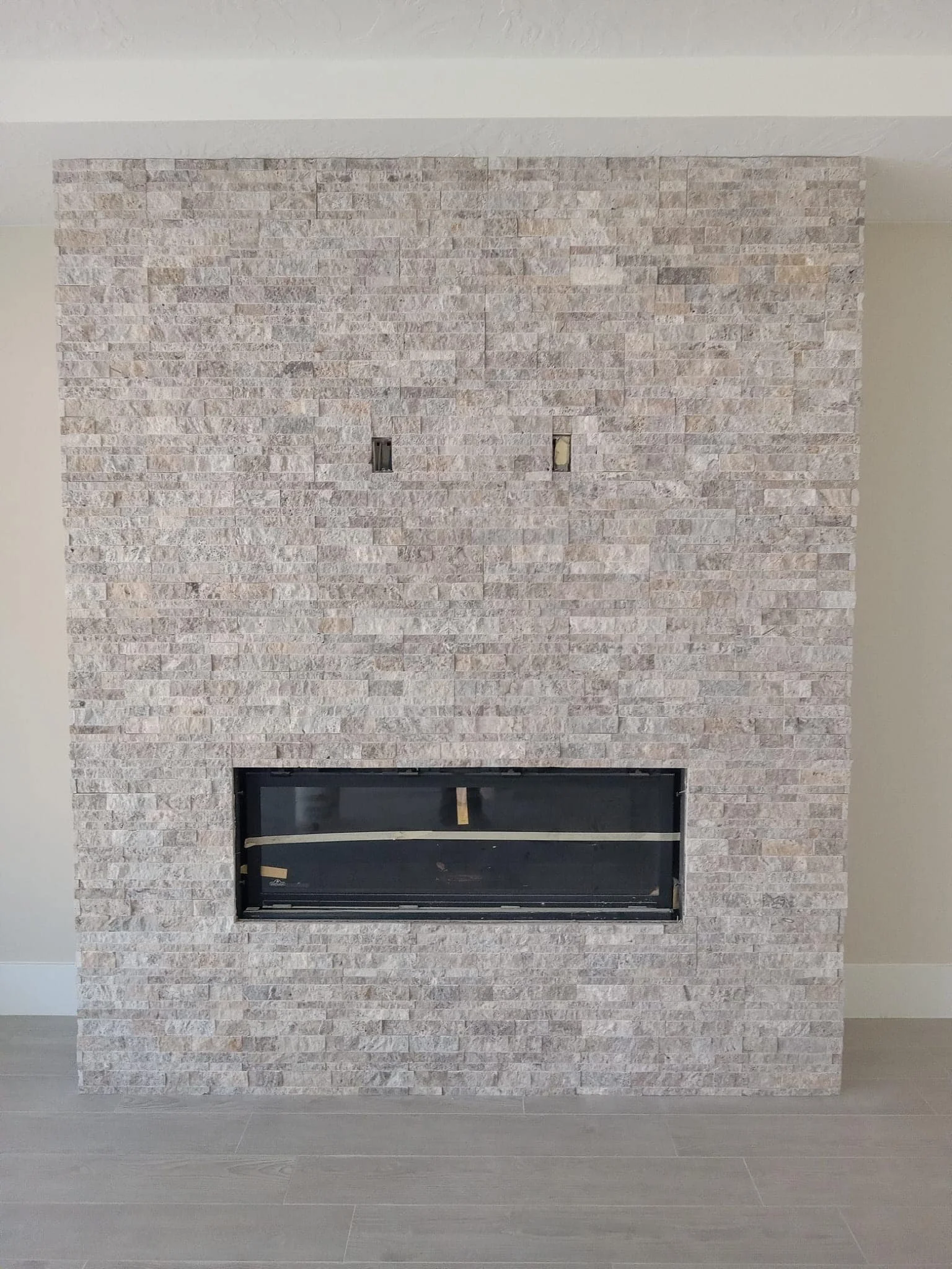 Interior view of a modern fireplace with a light-colored textured stone surround and a black opening at the bottom, set against a wall with light-colored paint and a gray wooden floor.
