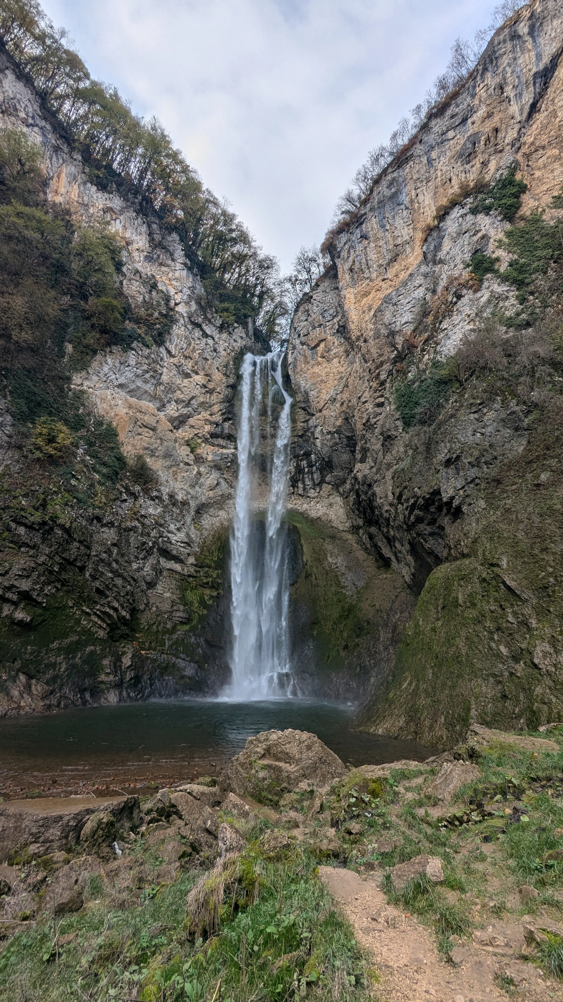 A waterfall cascading down a cliff into a pool below, surrounded by rocky terrain and sparse vegetation.