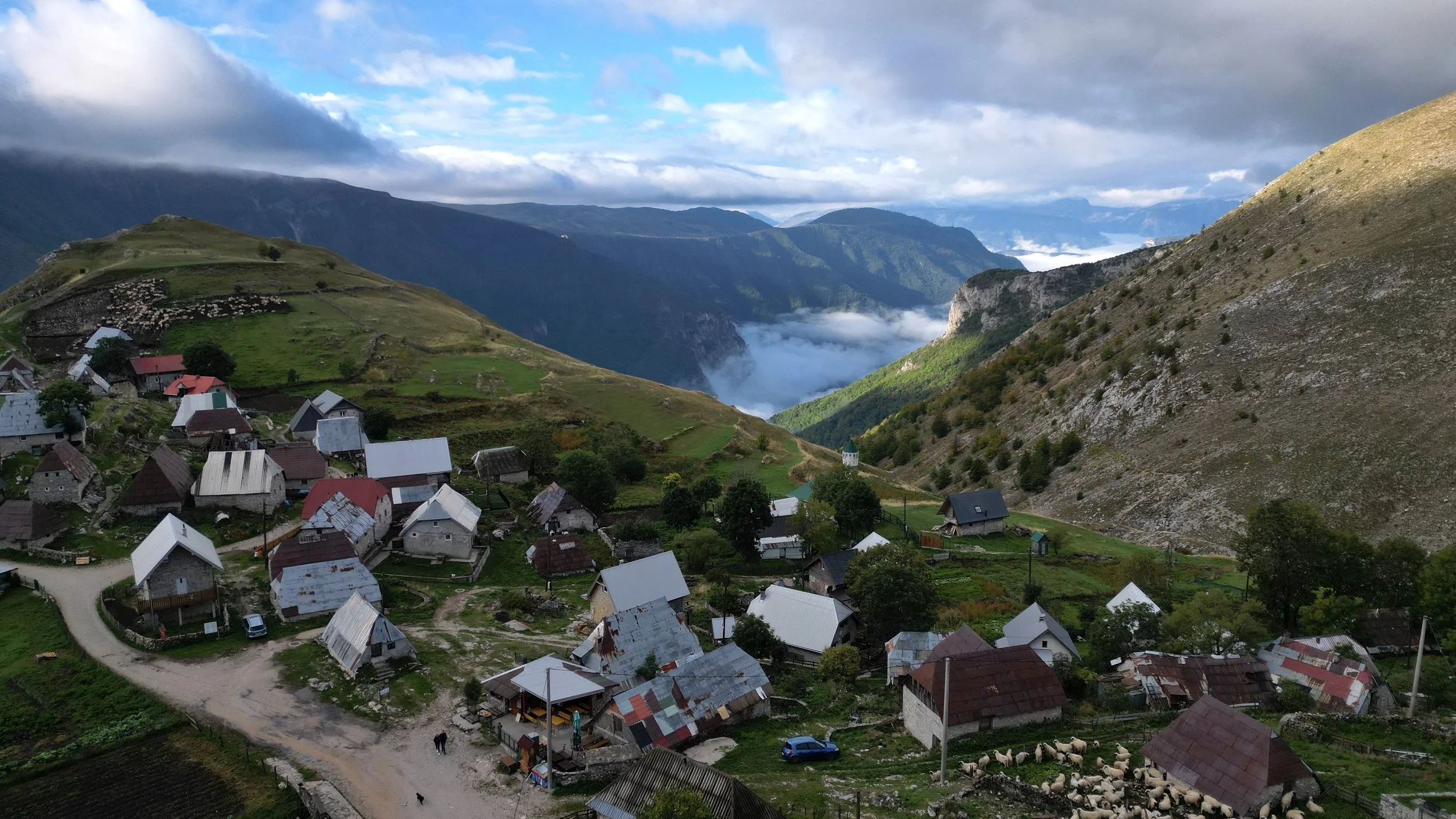 A mountain village with small houses with metal roofs, surrounded by green hills and dense forests, with mountains in the background and cloud-filled sky.