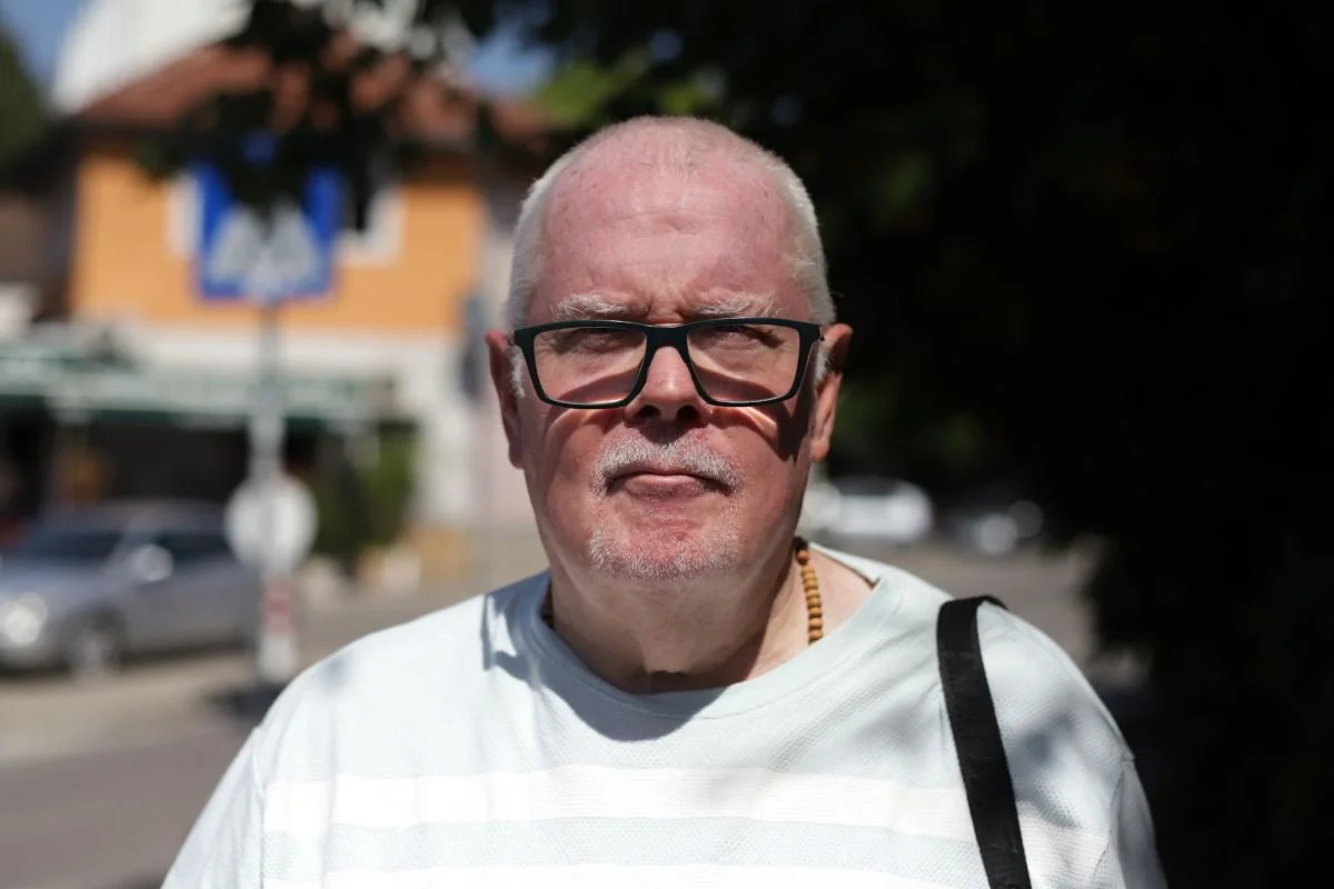 An older man with glasses and a beard standing outdoors in sunlight, wearing a white shirt and a beaded necklace.