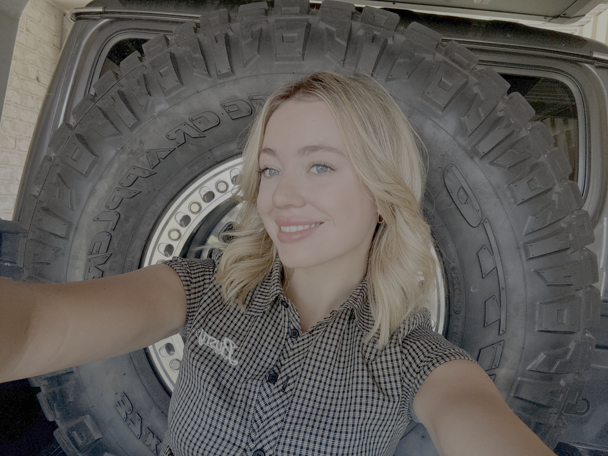 A young woman with light blonde hair and blue eyes taking a selfie in front of a large tire, possibly an off-road vehicle tire, with a brick wall in the background.