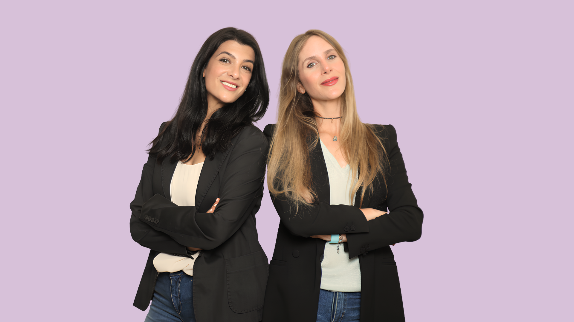 Two women standing with their arms crossed, smiling, and looking at the camera, against a lavender background.