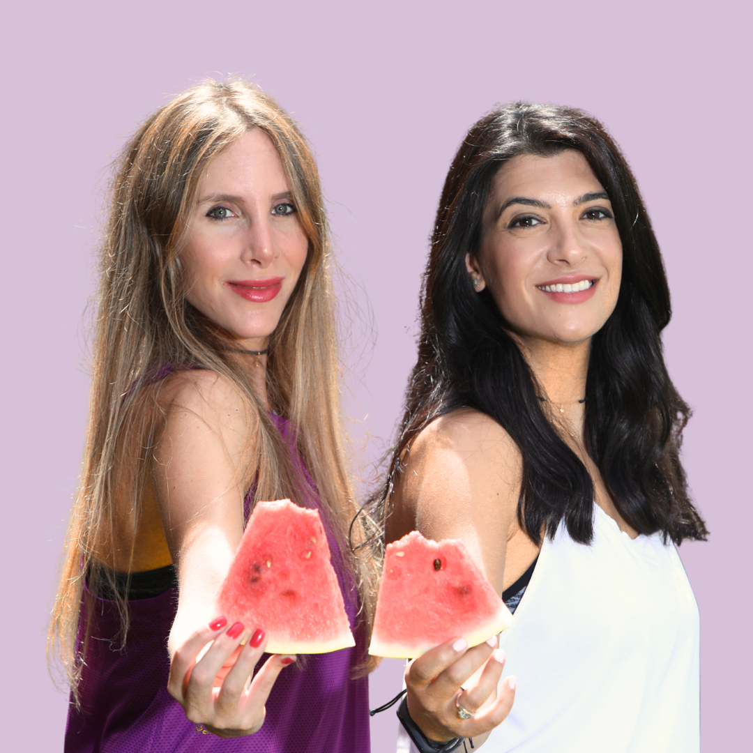 Two women with long hair, smiling, holding slices of watermelon, standing against a light purple background.