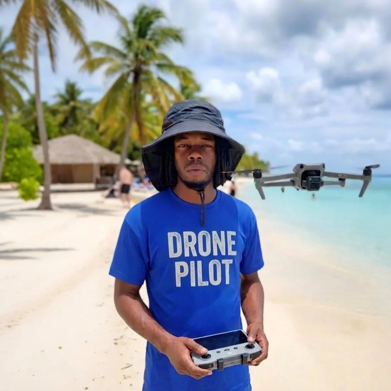 Man wearing a blue 'Drone Pilot' T-shirt and a wide-brimmed hat on a sandy beach with palm trees, holding a drone controller, while a drone flies nearby over clear water.