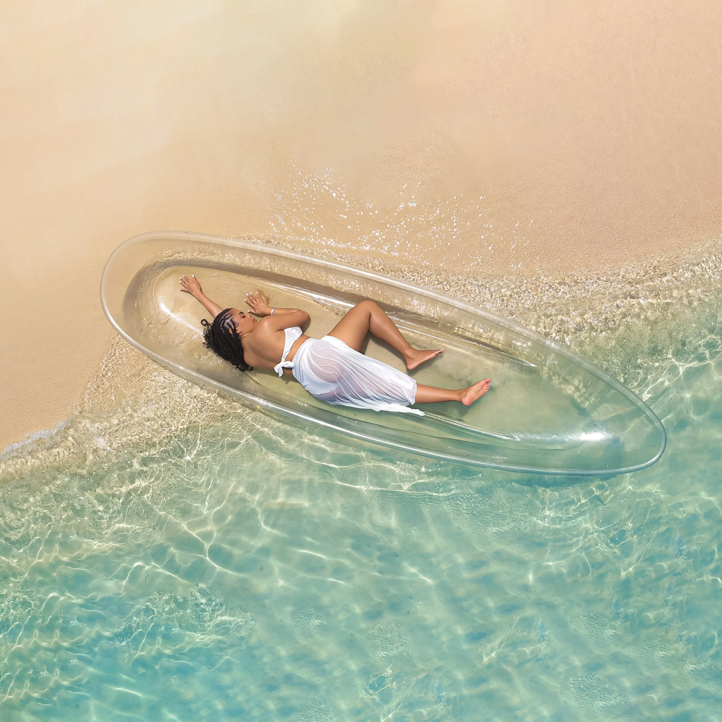 Person lying on a transparent water slide on a sandy beach, with the water below reflecting sunlight.