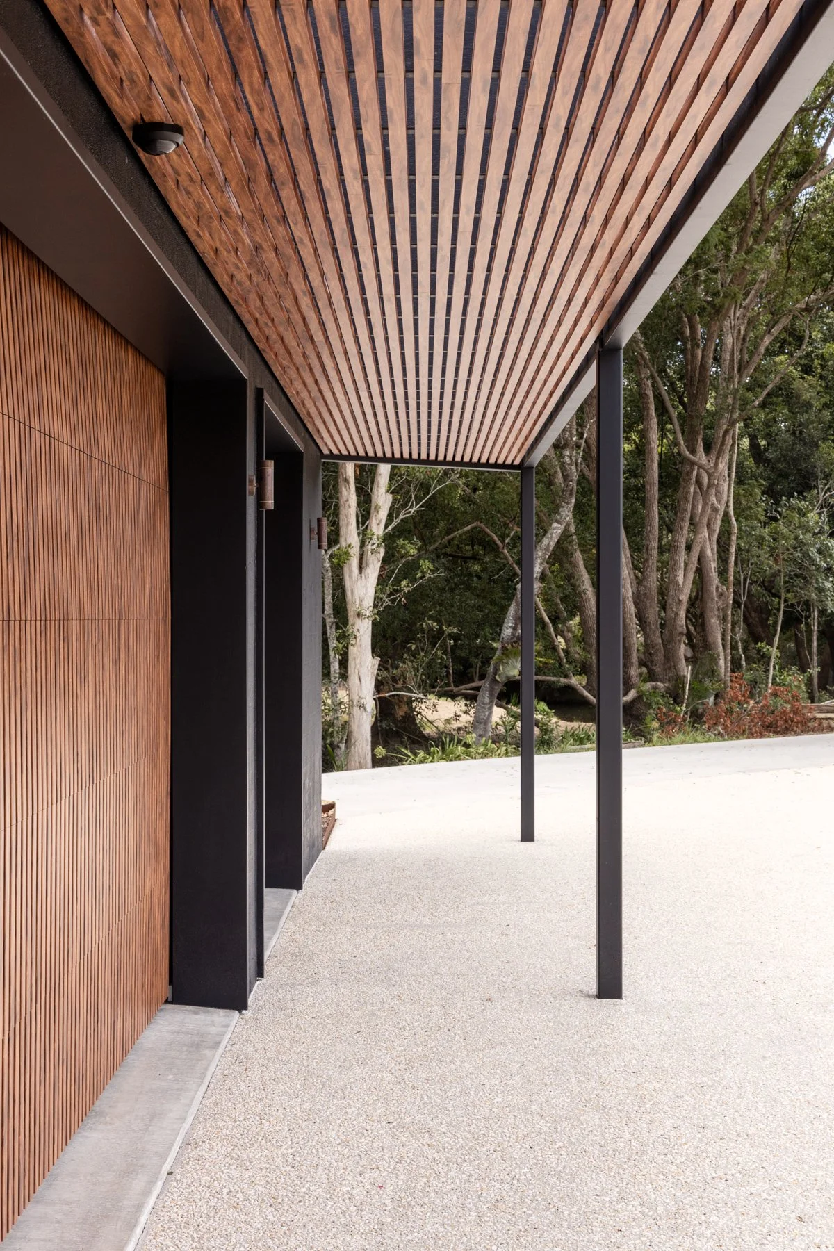 Modern outdoor porch with wooden slatted ceiling, black metal supports, and a textured concrete floor. Background features trees and natural surroundings.