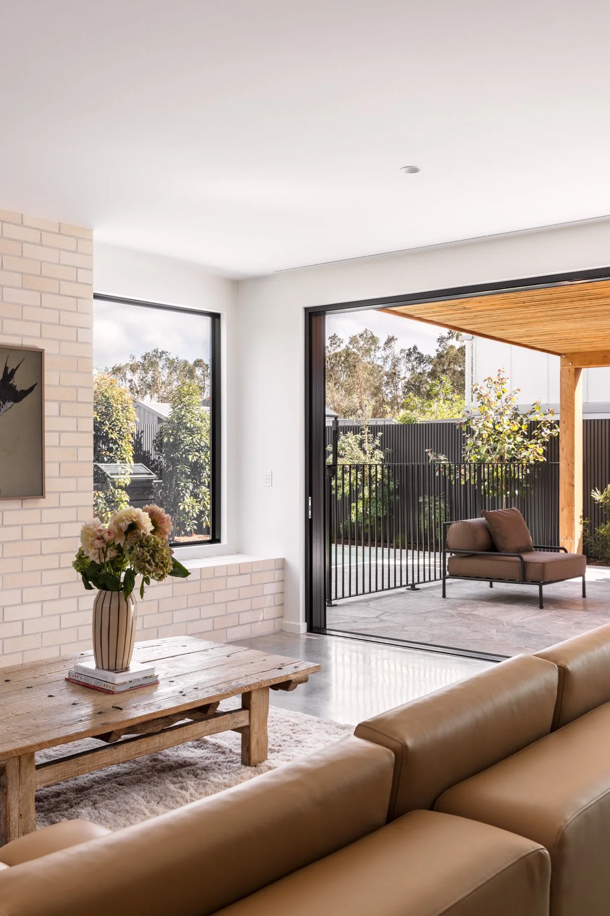Modern living room with beige leather sofa, wooden coffee table with a vase of pink and cream flowers, large window, and sliding glass door leading to an outdoor patio with a cushioned outdoor chair and greenery.
