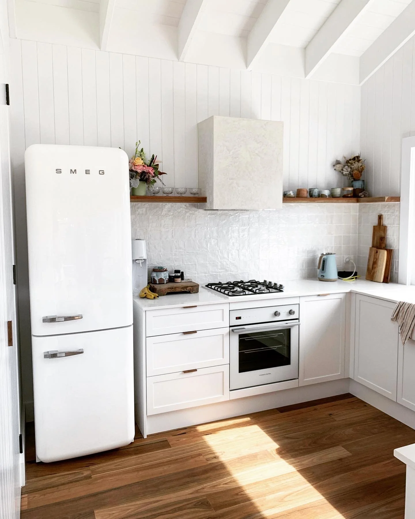 A white kitchen with wooden accents, featuring a vintage SMEG refrigerator, a white oven, a gas stove, and a countertop with a kettle, cutting boards, and small jars. Wooden floating shelves hold vases and ceramics, and there are dried flowers on the shelves.