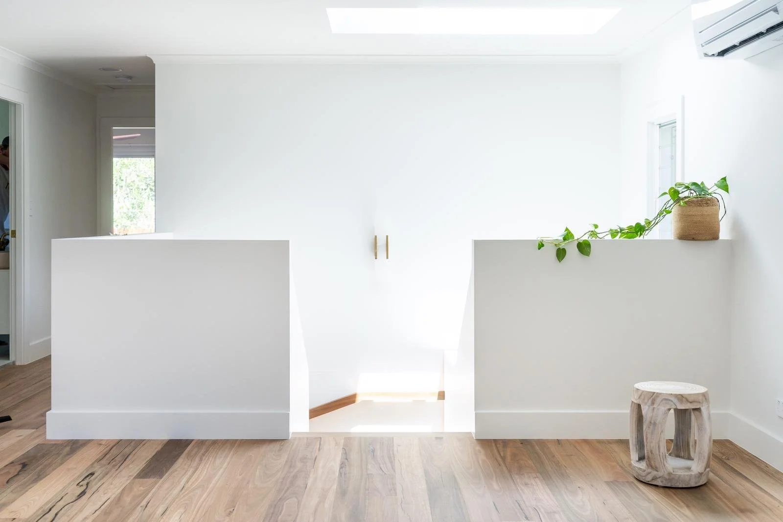 Minimalist white interior with a small staircase, a potted plant, a wooden stool, and natural light from a window.