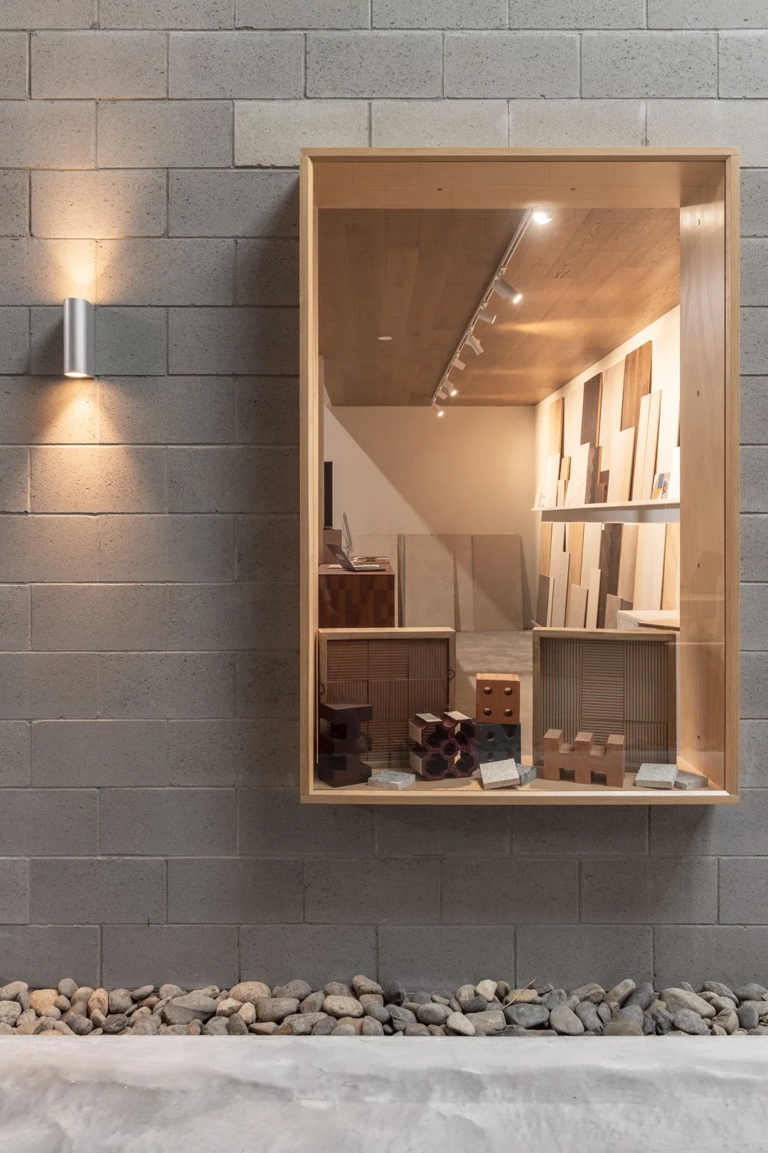 View through a glass display case showing wood samples and building materials inside an interior design showroom. The case is mounted on a concrete brick wall outside.