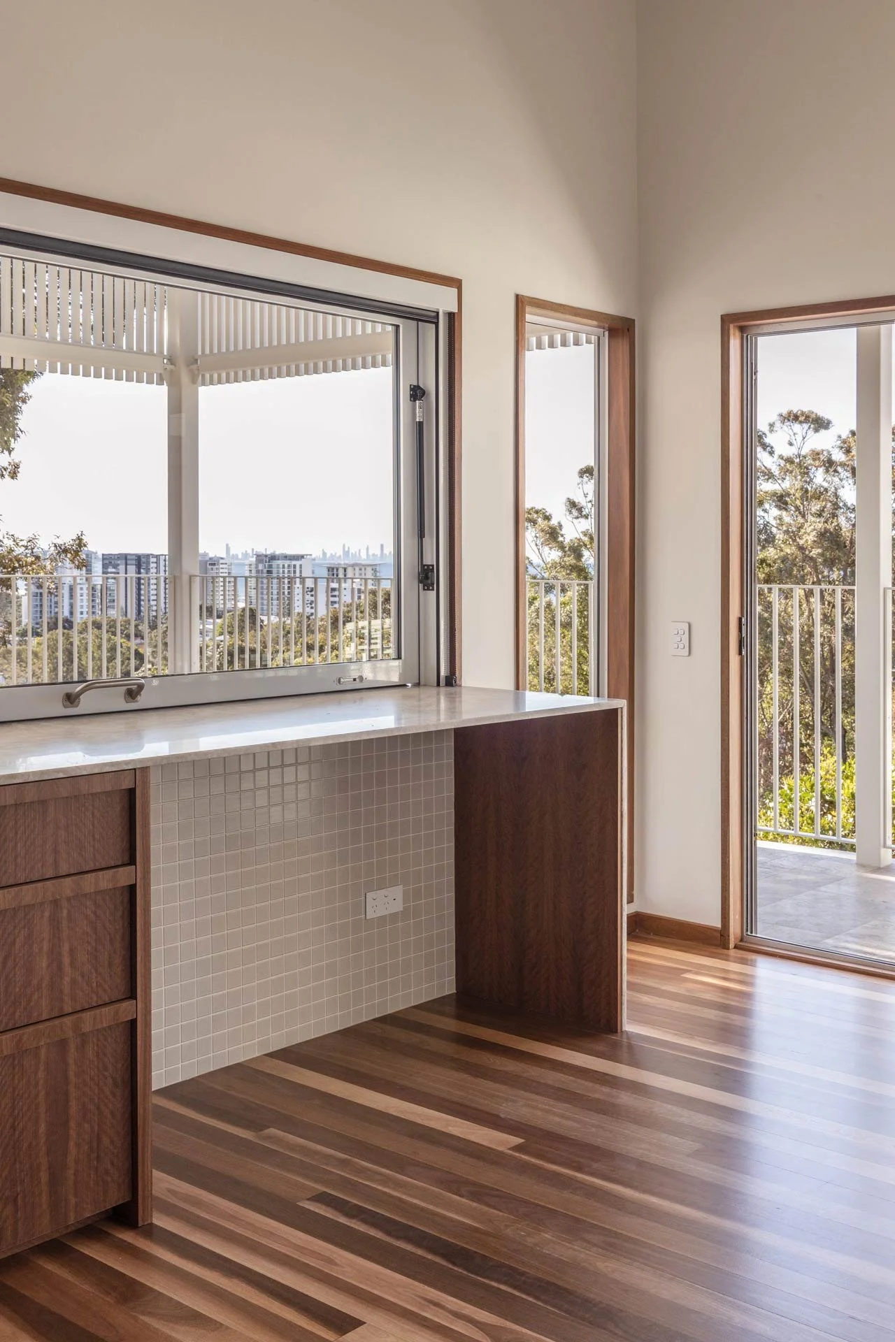 Interior view of a room with large windows, wooden flooring, and a balcony with a view of city buildings and trees.