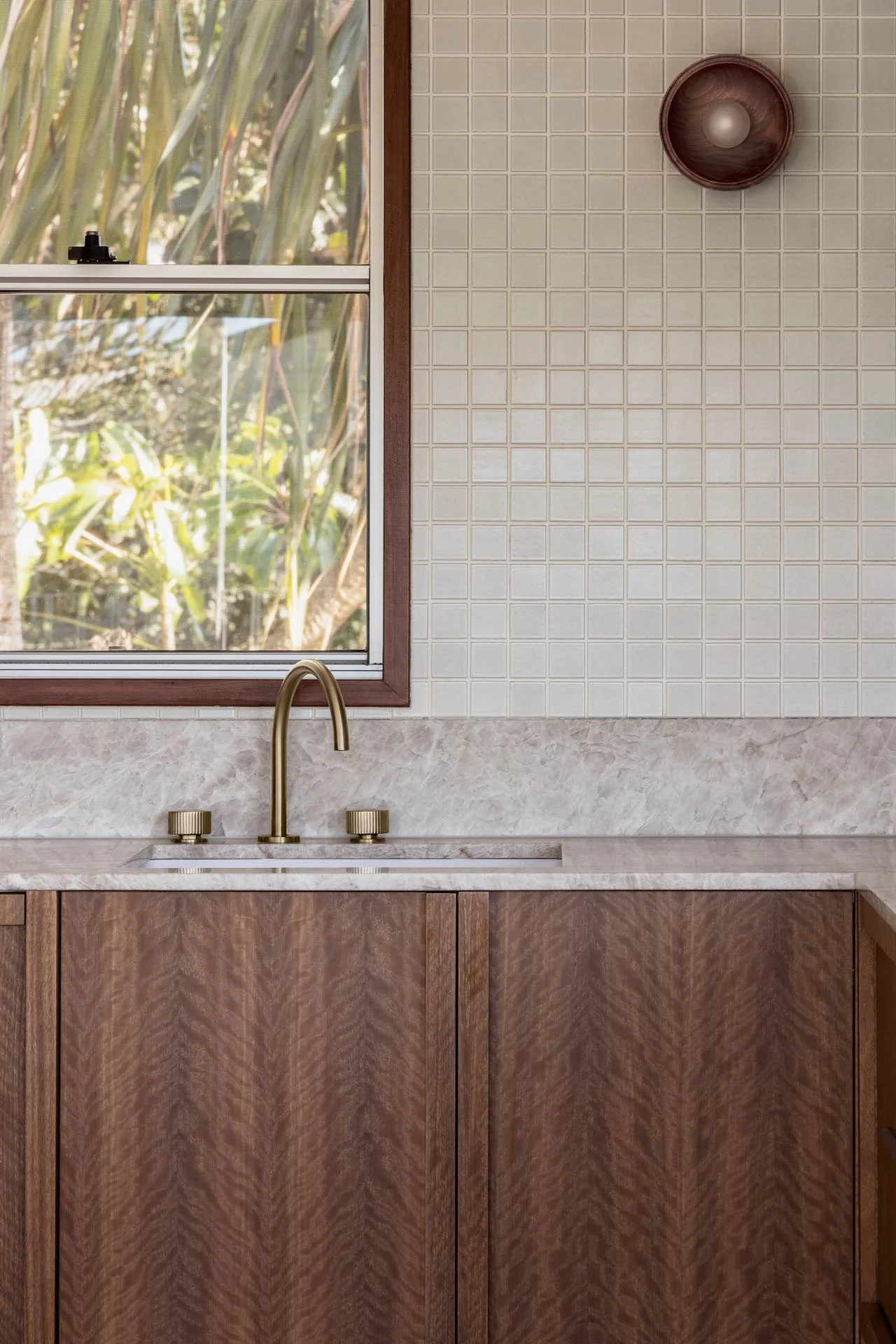 Interior view of a kitchen sink with a gold faucet, wooden cabinets, a window showing outdoor greenery, beige tiled wall with a round wooden shelf and a light fixture.