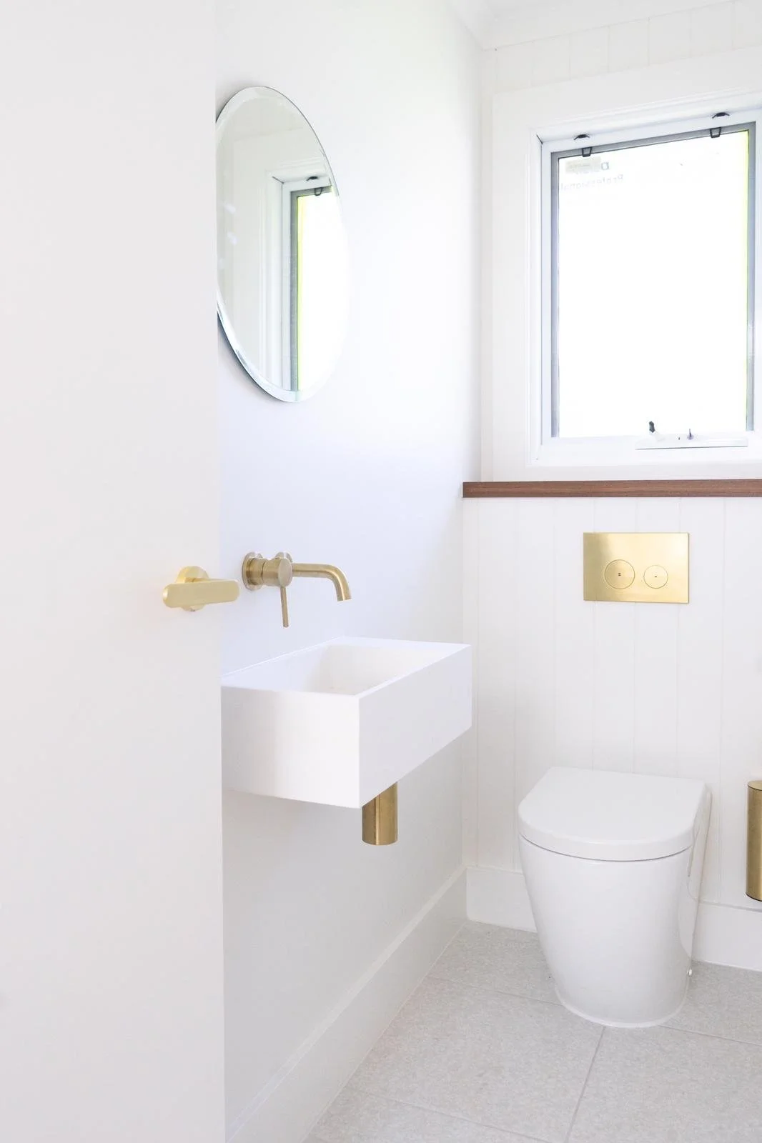 A minimalist white bathroom with a small rectangular sink, a wall-mounted gold faucet, a white toilet, a round mirror, a window, and gold fixtures.