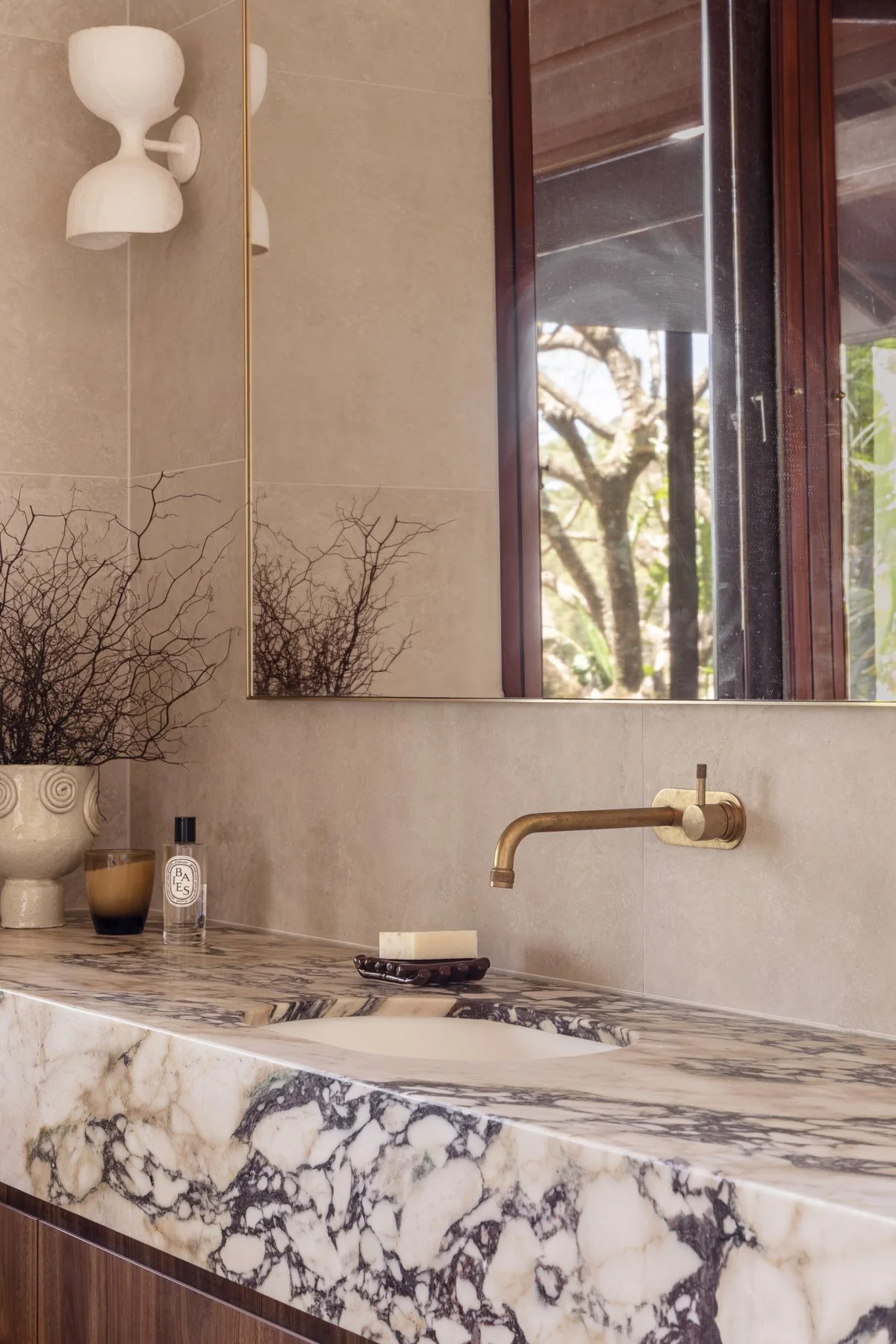 A bathroom with a marble countertop, a brass faucet, a large mirror, and a window reflecting trees outside. Decor includes a vase with twigs, candles, and perfume bottles.