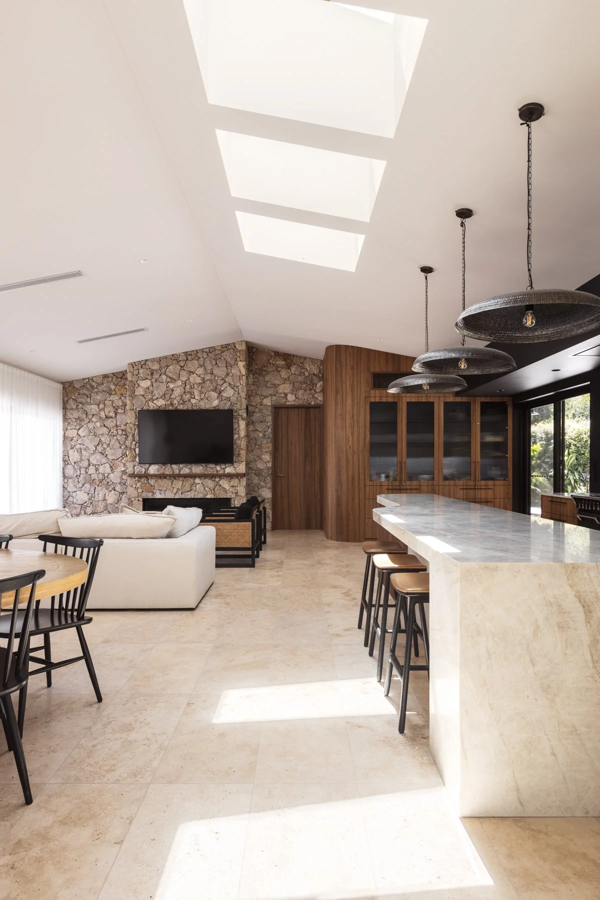 Modern open-concept kitchen and living room with stone accent wall, mounted flat-screen TV, white sofa, bar seating, wood cabinetry, and skylights.