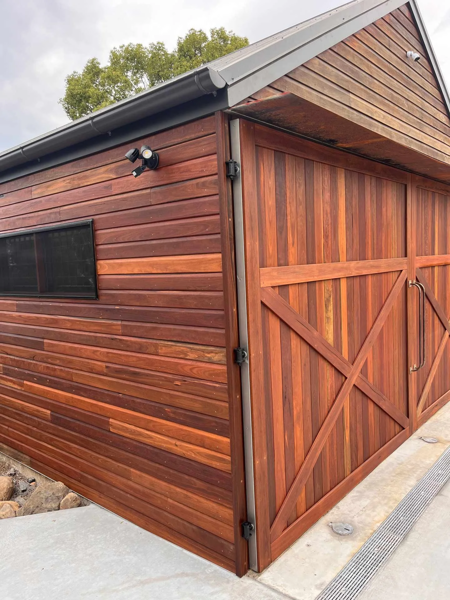 A wooden shed with a sliding barn door, a small window, security cameras, and outdoor lighting.