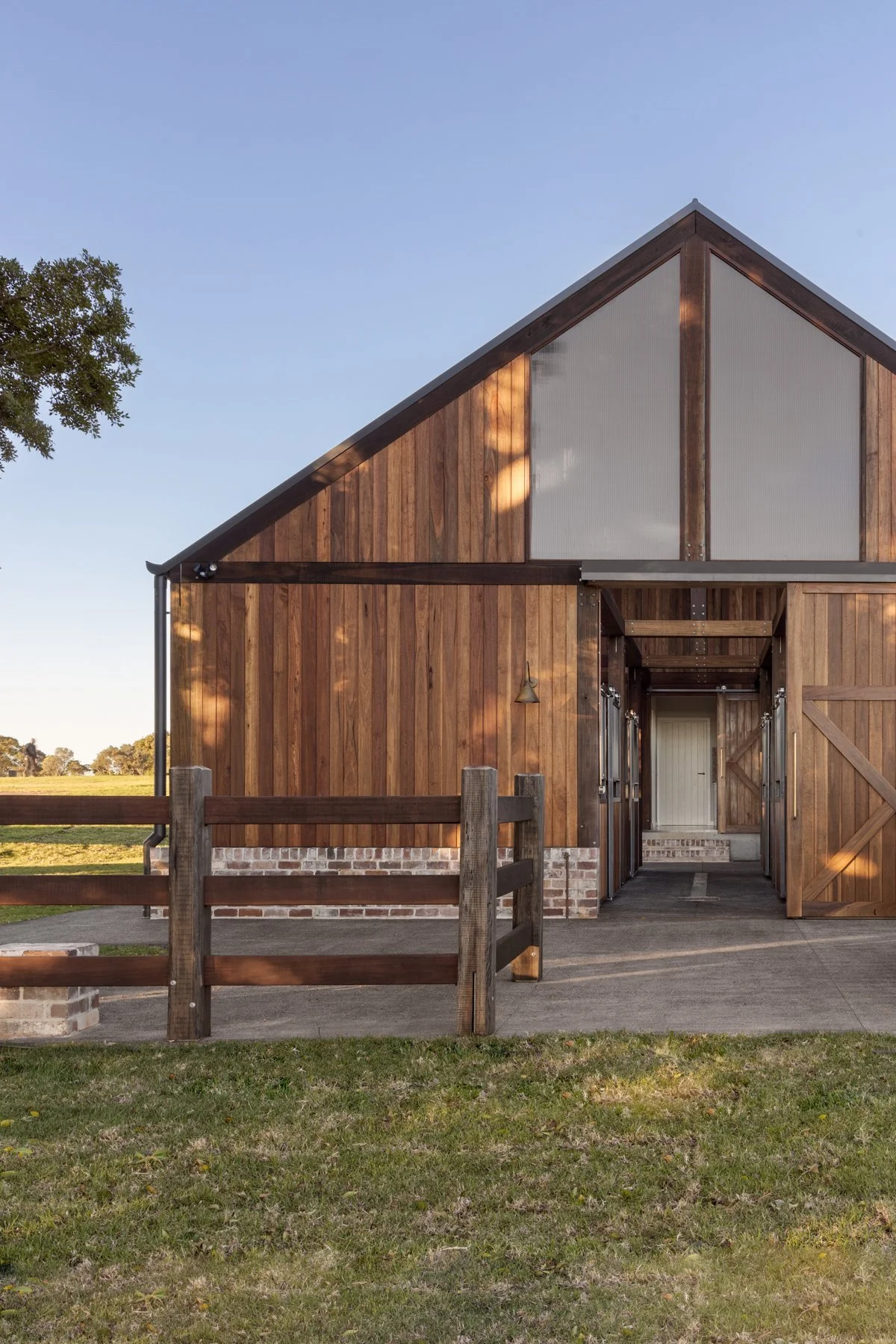 A rustic barn with wooden siding and brick foundation, with open sliding doors revealing an interior passage. There is a wooden fence in front, and a grassy area with trees and a clear blue sky in the background.