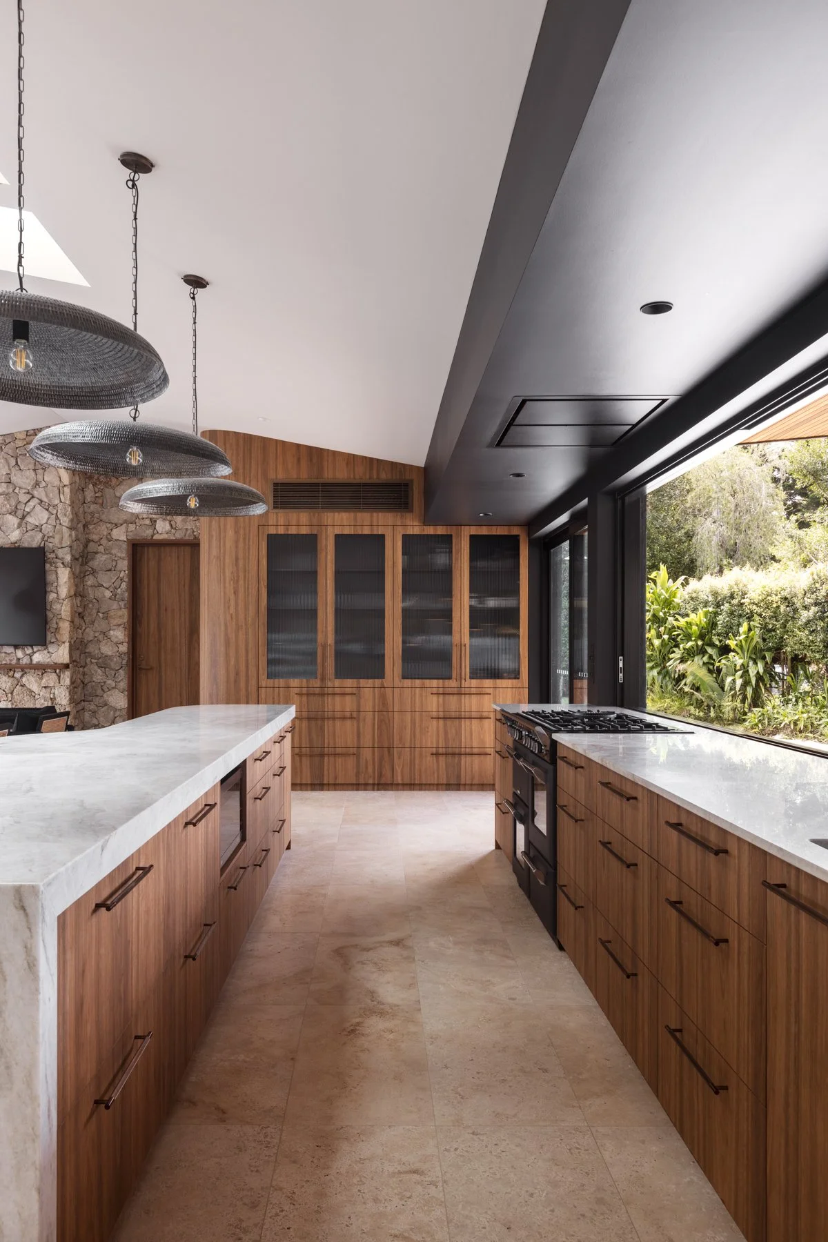 Modern kitchen featuring wooden cabinets, a white marble island, large window with outdoor view, black stove, stone accent wall, and minimalist lighting fixtures.