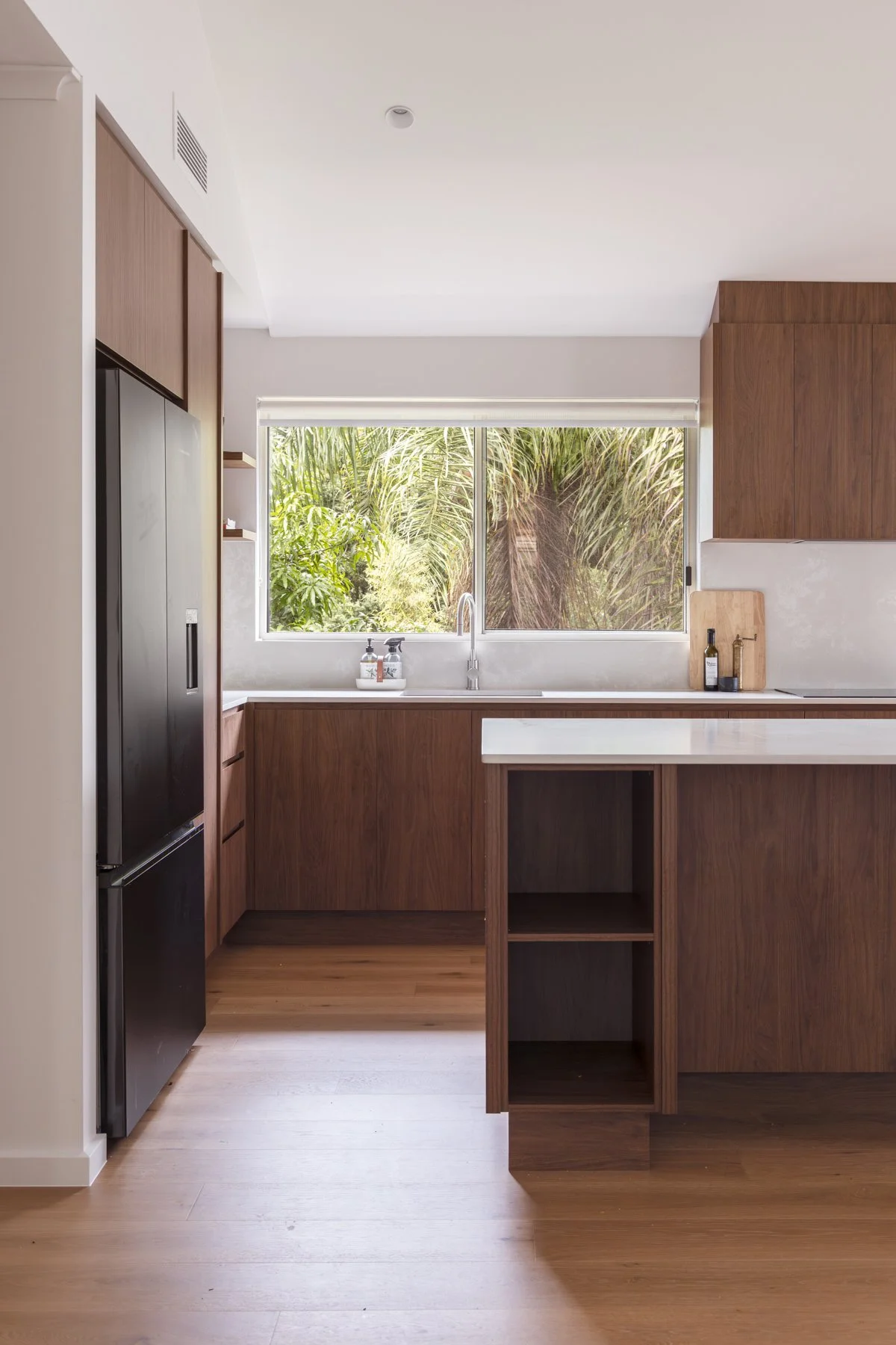 Modern kitchen with wooden cabinets, large window showing greenery outside, and a white countertop with bottles and a cutting board.
