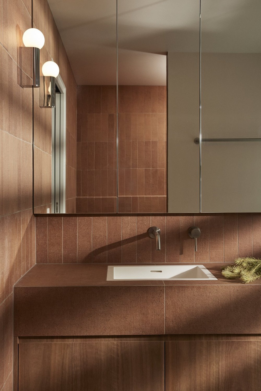 A modern bathroom with reddish-brown tiled walls, a white rectangular sink, dual modern faucets, a mirrored cabinet, and wall-mounted globe lights.
