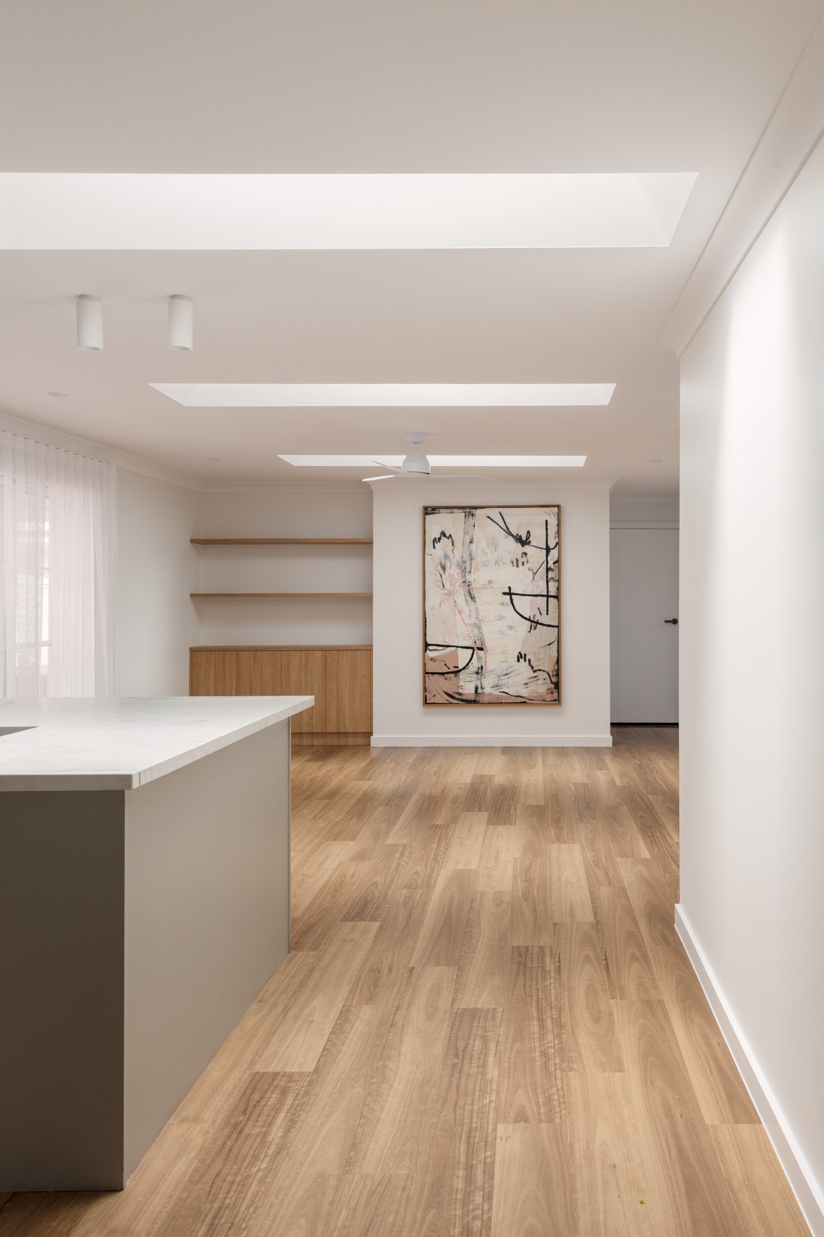 Modern dining area with light wood flooring, white walls, a white kitchen island, wooden open shelves, and abstract artwork on the wall.