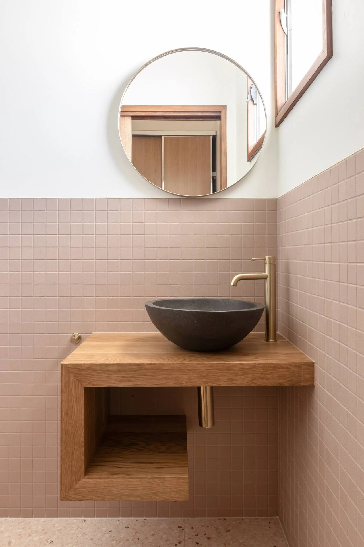 A modern bathroom sink area with a round mirror, a black stone vessel sink, a gold faucet, a wooden vanity, pink tiled walls, a wooden window frame, and a beige tiled floor.