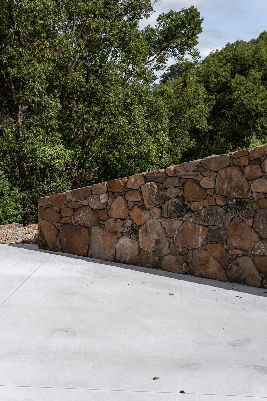 A stone wall with large, irregularly shaped rocks, next to a smooth, white concrete surface, and green trees in the background under a partly cloudy sky.