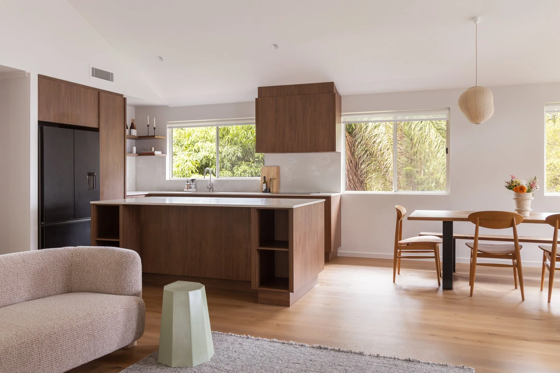 Open-concept kitchen and dining area with wooden furniture, a black refrigerator, white walls, large windows with greenery outside, a beige sofa, a small white stool, and a dining table with chairs and a flower arrangement.