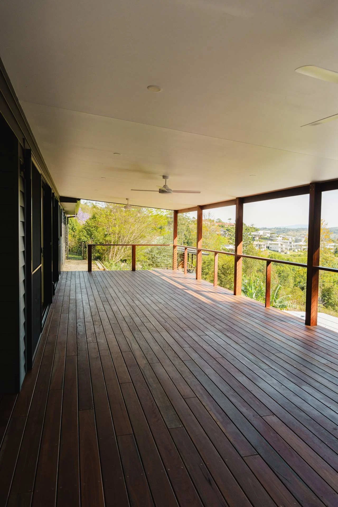 Empty wooden deck with a wooden railing, ceiling fans, and a view of greenery and distant houses.