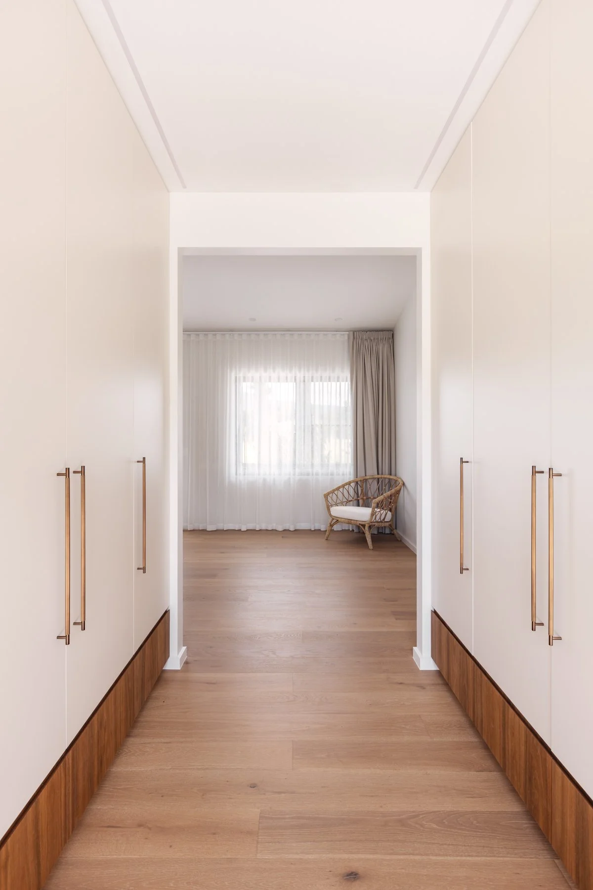 A walk-in closet with white cabinets and wooden floors, leading to a room with a large window and beige curtains, and a wicker chair with a white cushion.