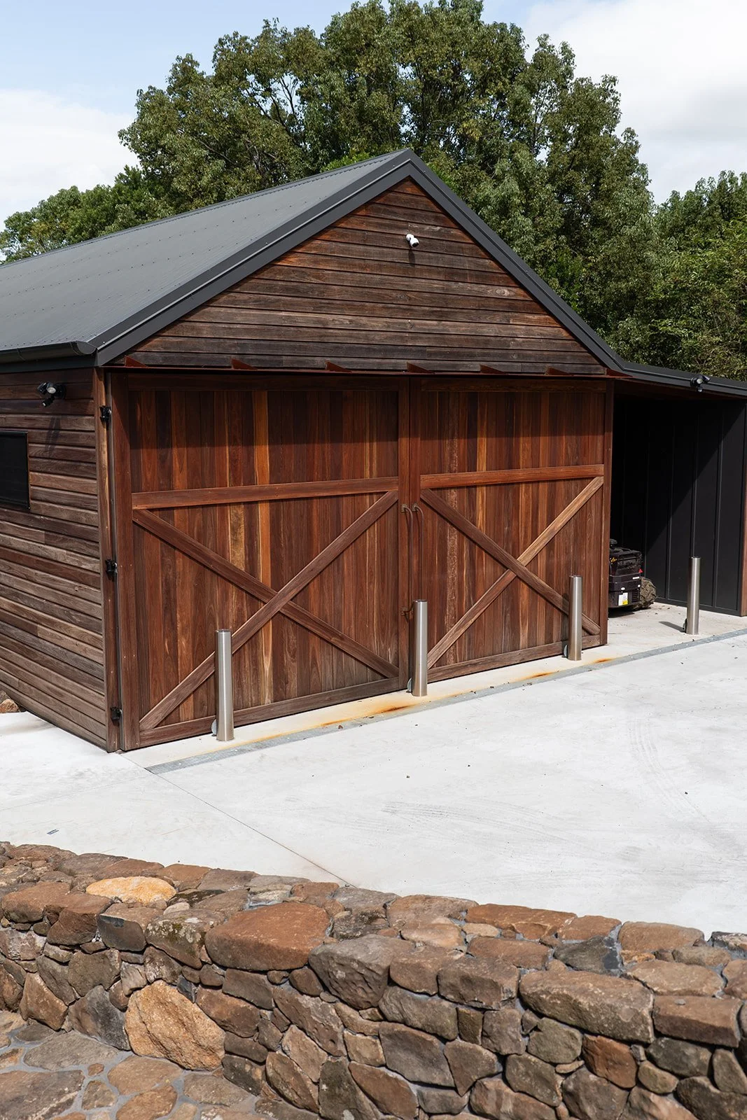 A wooden garage with double doors, a metal roof, and security bollards in front, surrounded by a stone wall and trees in the background.