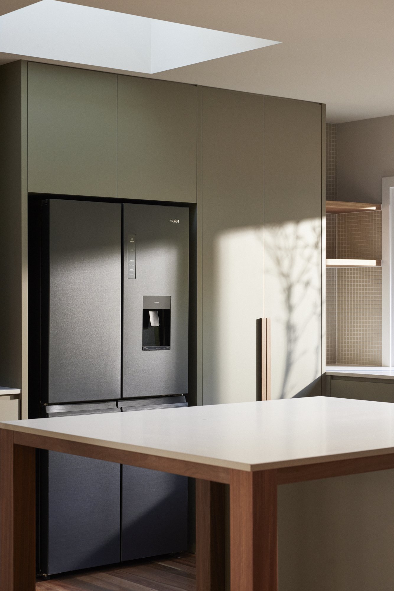 A modern kitchen with a black refrigerator, beige cabinets, and a wooden island with a white countertop. Sunlight casts shadows on the wall.