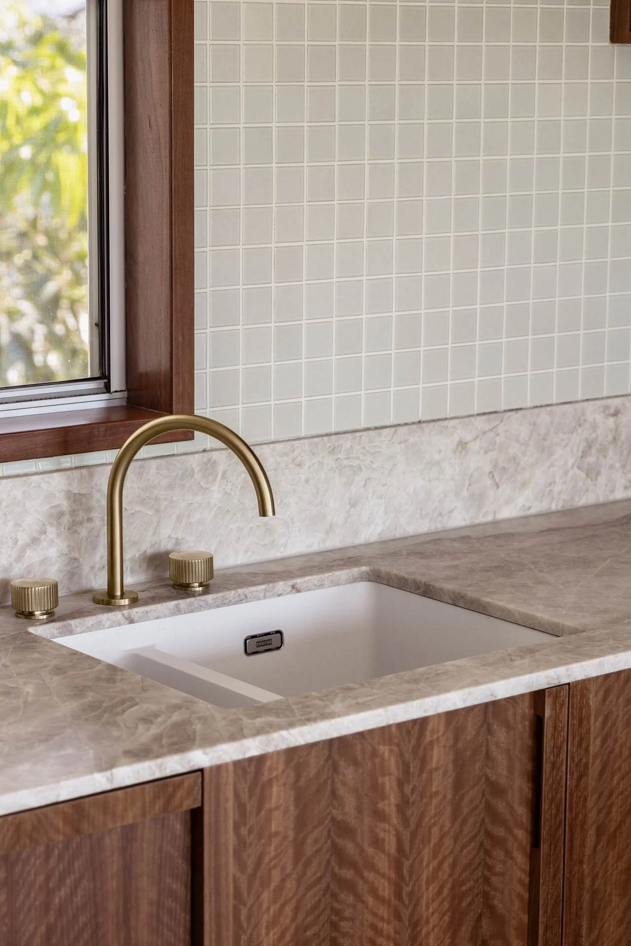 A kitchen sink area with a white undermount sink, a gold-colored faucet, and two matching knobs. The countertop is a beige marble with wood cabinetry underneath. Behind the sink, the wall is tiled with small, cream-colored square tiles, and there is a window with a wooden frame to the left.