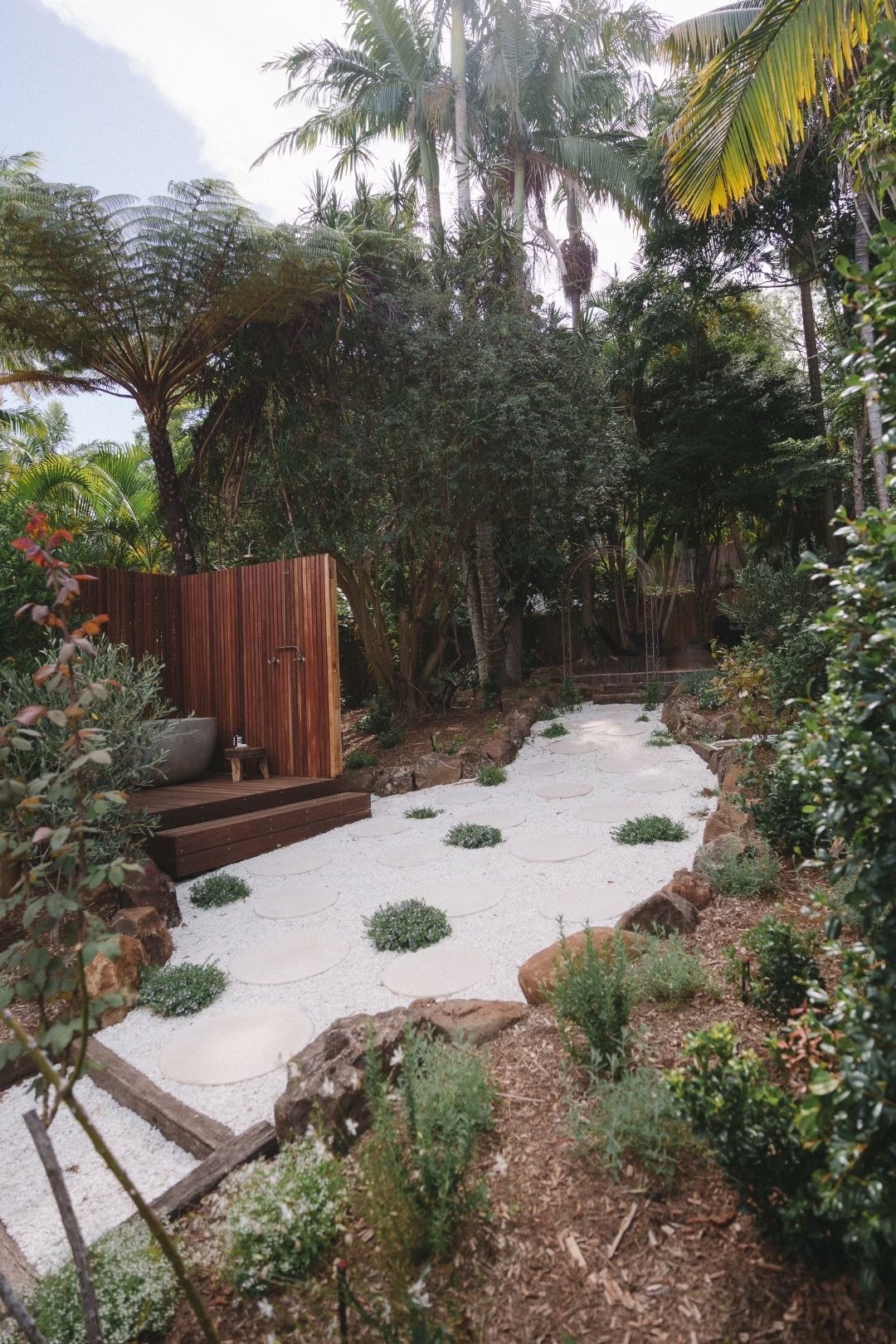 A lush garden with tropical trees and plants, a wooden privacy fence, and a peaceful white pebble pathway bordered by rocks and greenery.