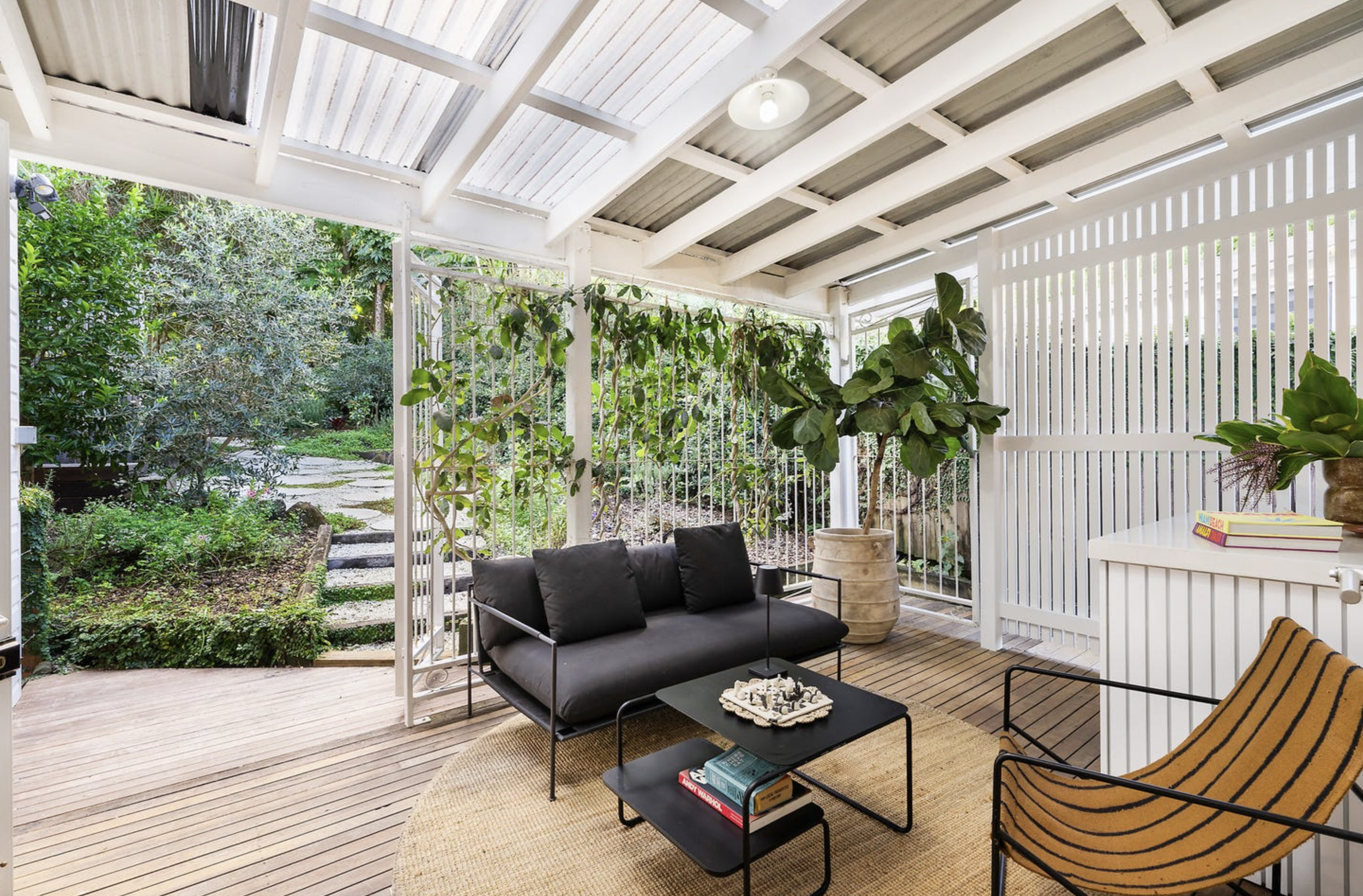 Back porch with black outdoor sofa, coffee table, green plants, and a garden view with trees and shrubs.