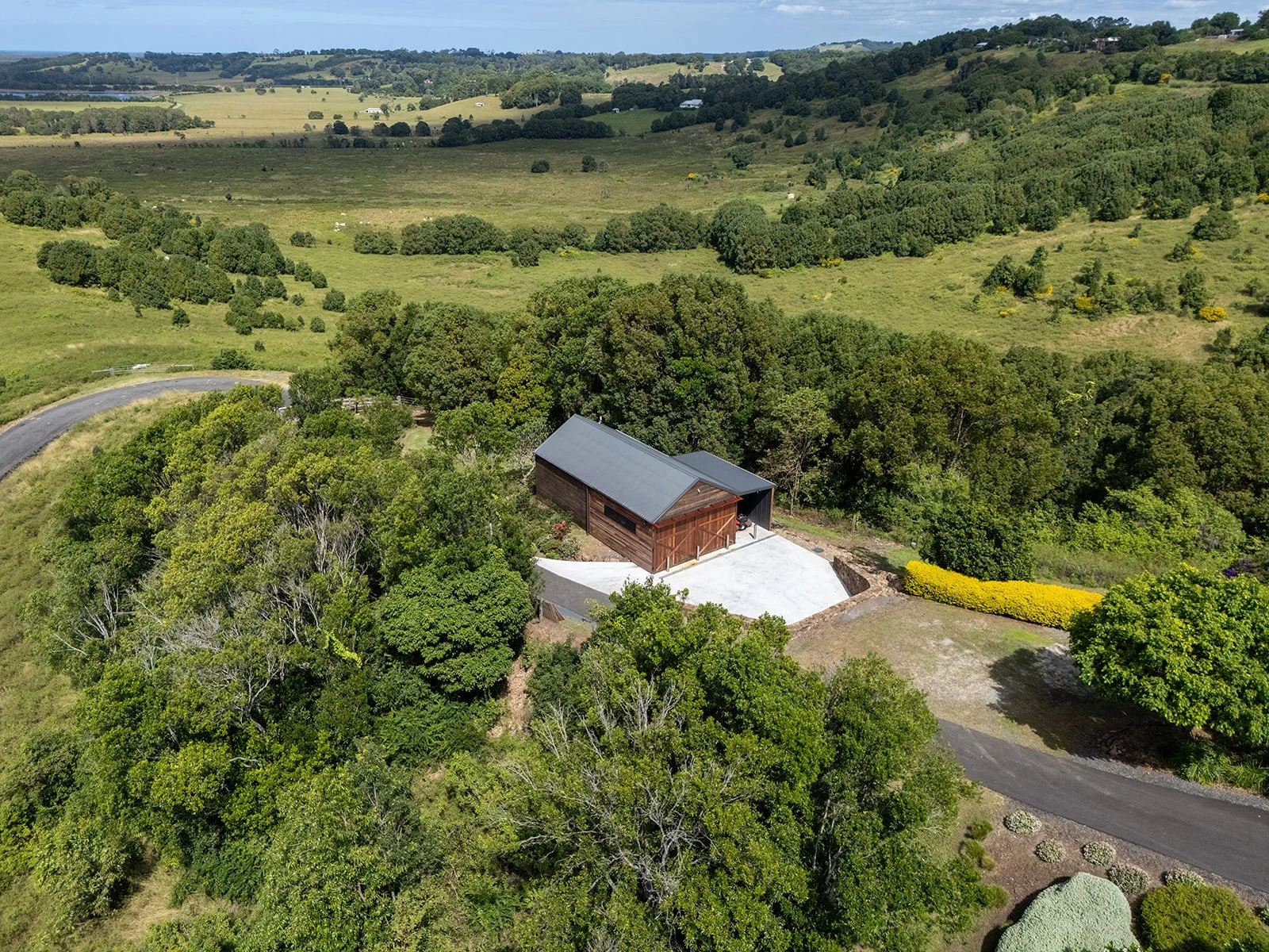 A wooden barn with a metal roof surrounded by lush green trees in a rural landscape, with rolling hills and fields in the background.