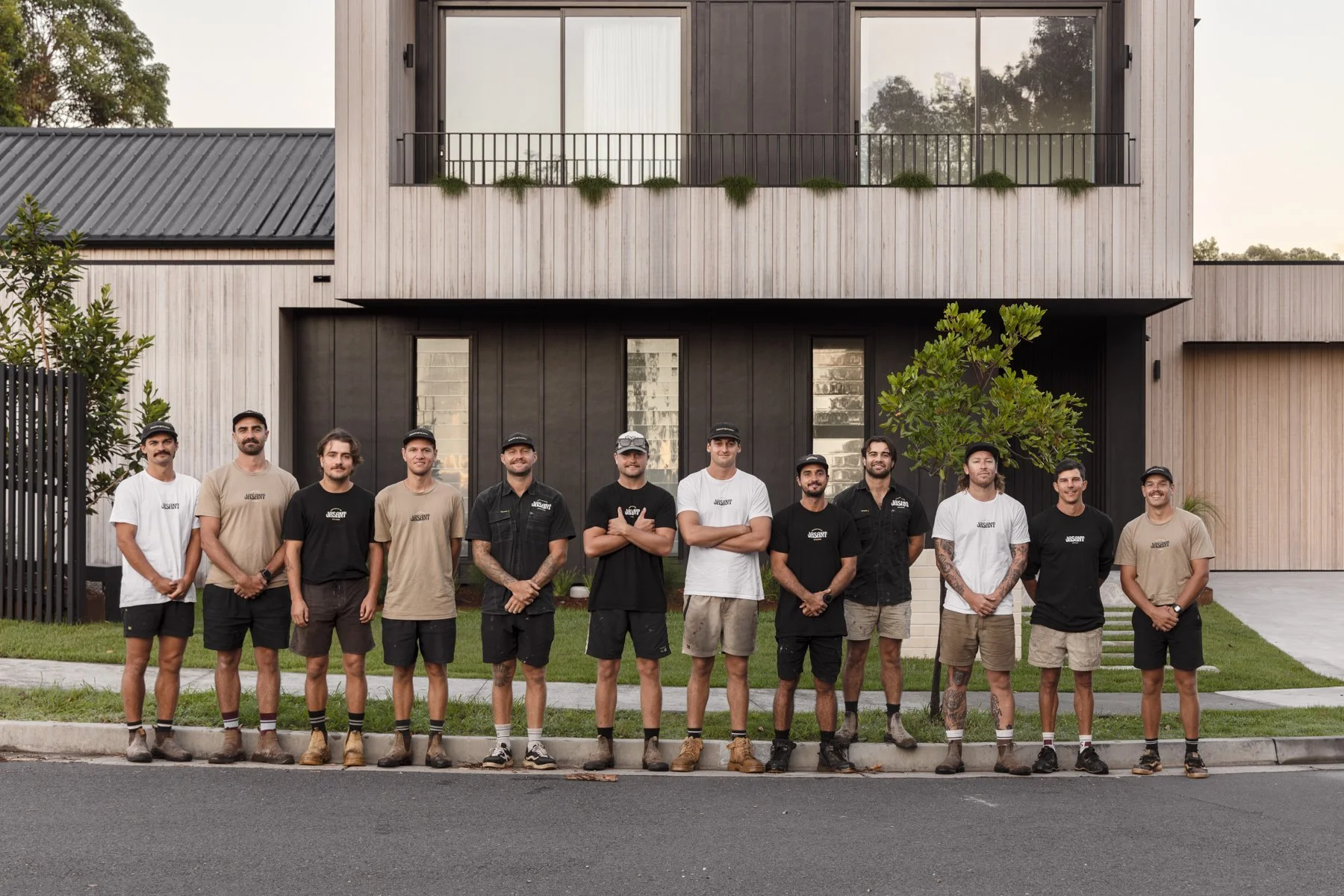 A group of eleven construction workers standing in front of a modern two-story house with wooden and black paneling. They are outdoors on the sidewalk, wearing work attire and smiling at the camera.