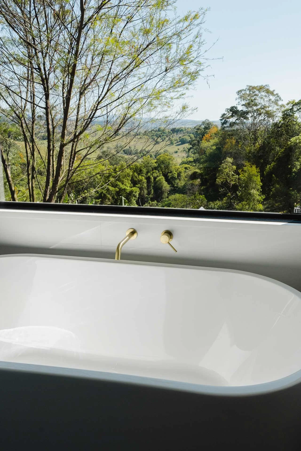 View of a spacious window overlooking a lush green landscape with trees, hills, and a clear blue sky, seen from inside a modern bathtub with a gold-colored faucet.