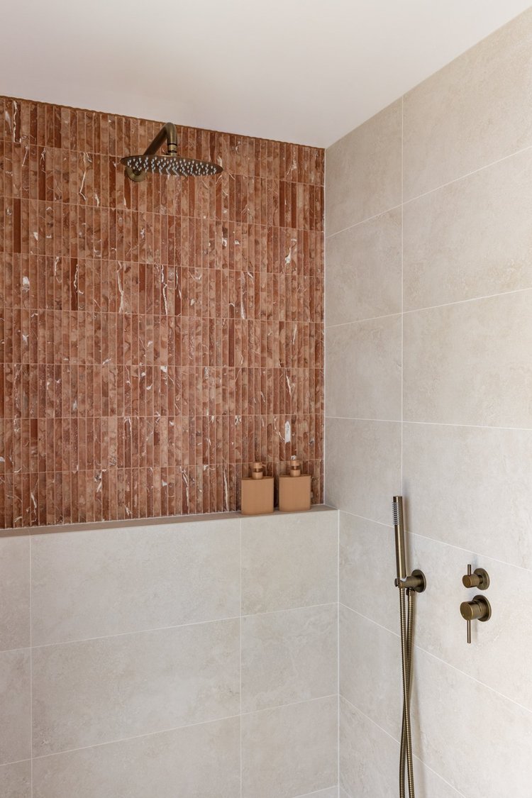 A modern shower with beige tiles on the lower half and red-brown textured tiles on the upper half, featuring a round overhead showerhead, a handheld shower wand with a hose, and three beige containers with candles placed on a ledge.