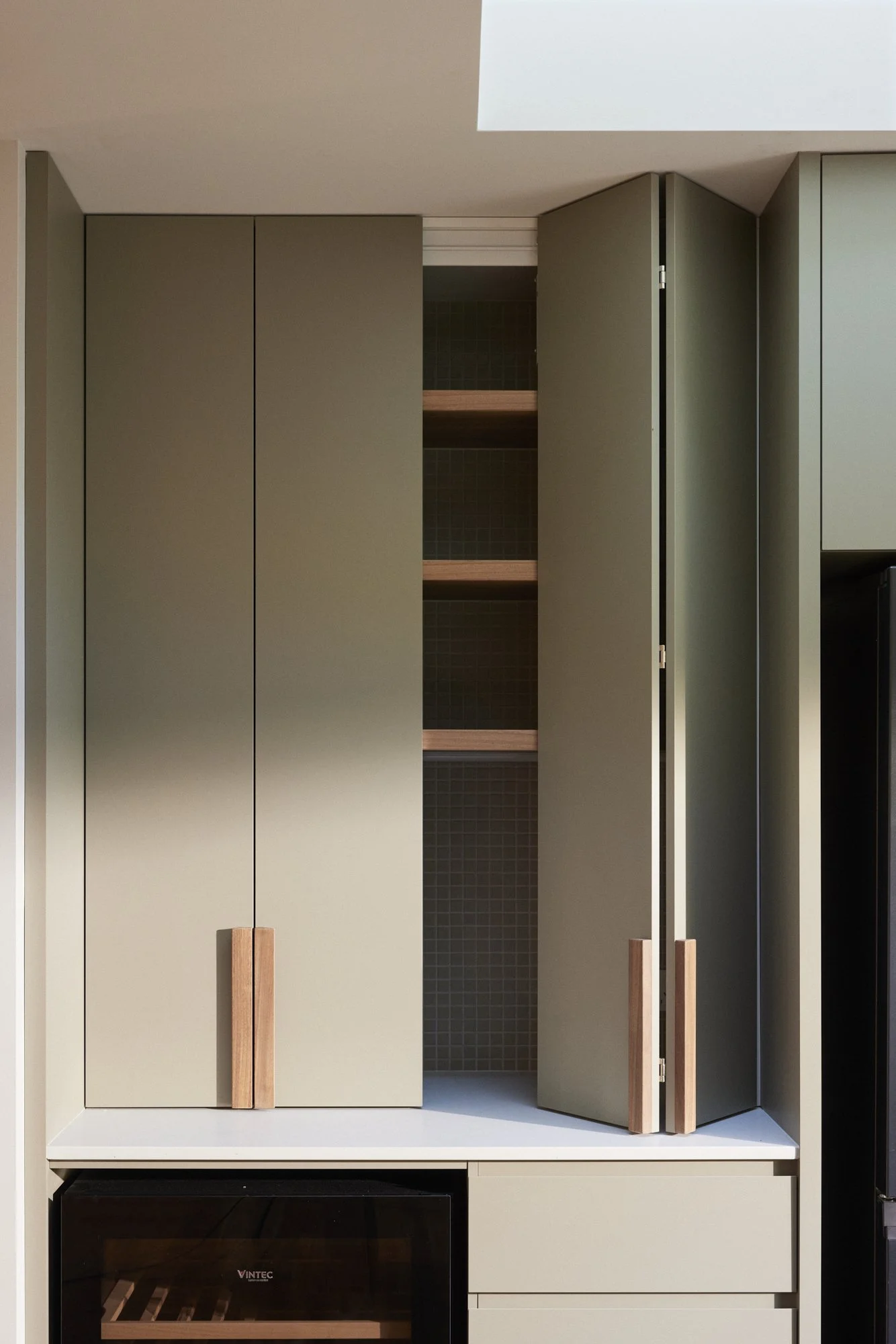 A modern kitchen cabinet with closed green doors and an open shelf in the middle. Next to the cabinet, on the right, is a door with wooden handles, and beneath the cabinets is a built-in oven with a wooden wine rack inside.